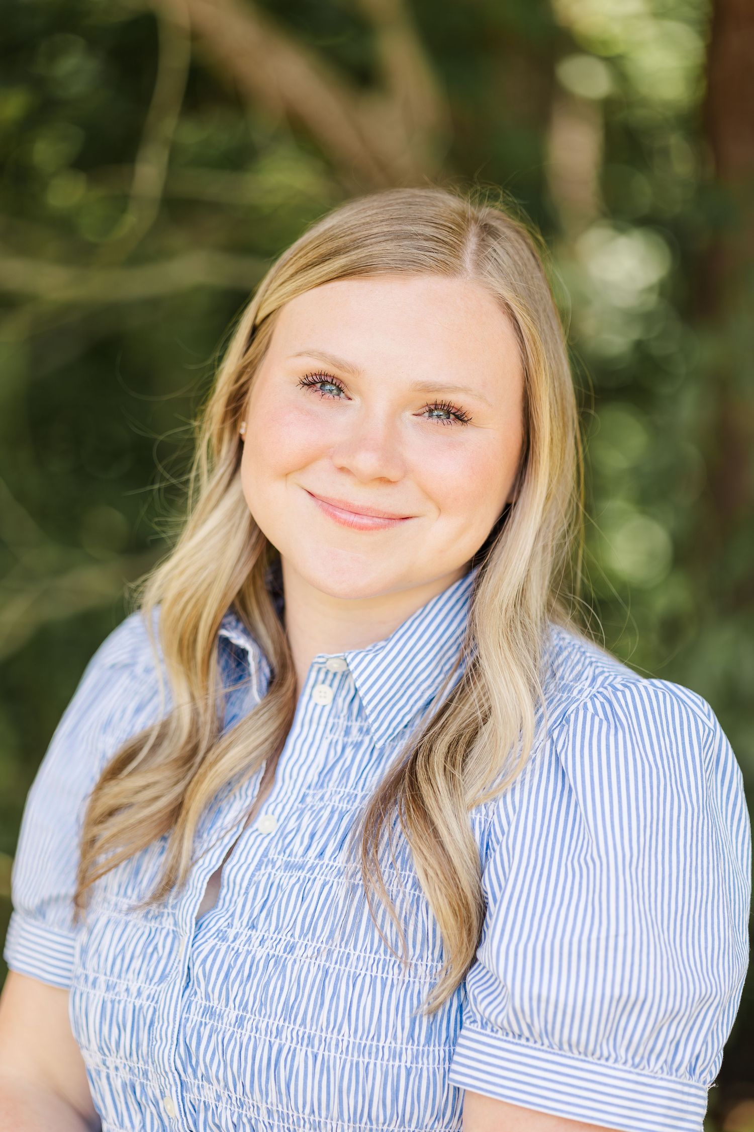 Smiling young blonde woman in blue striped shirt poses outdoors with green trees in background.