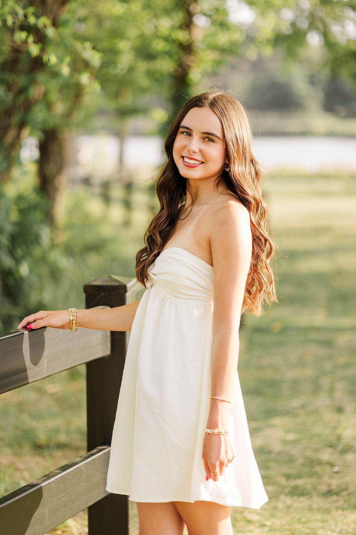 Young woman in white strapless mini dress smiling outdoors by a wooden fence in a lush green park.