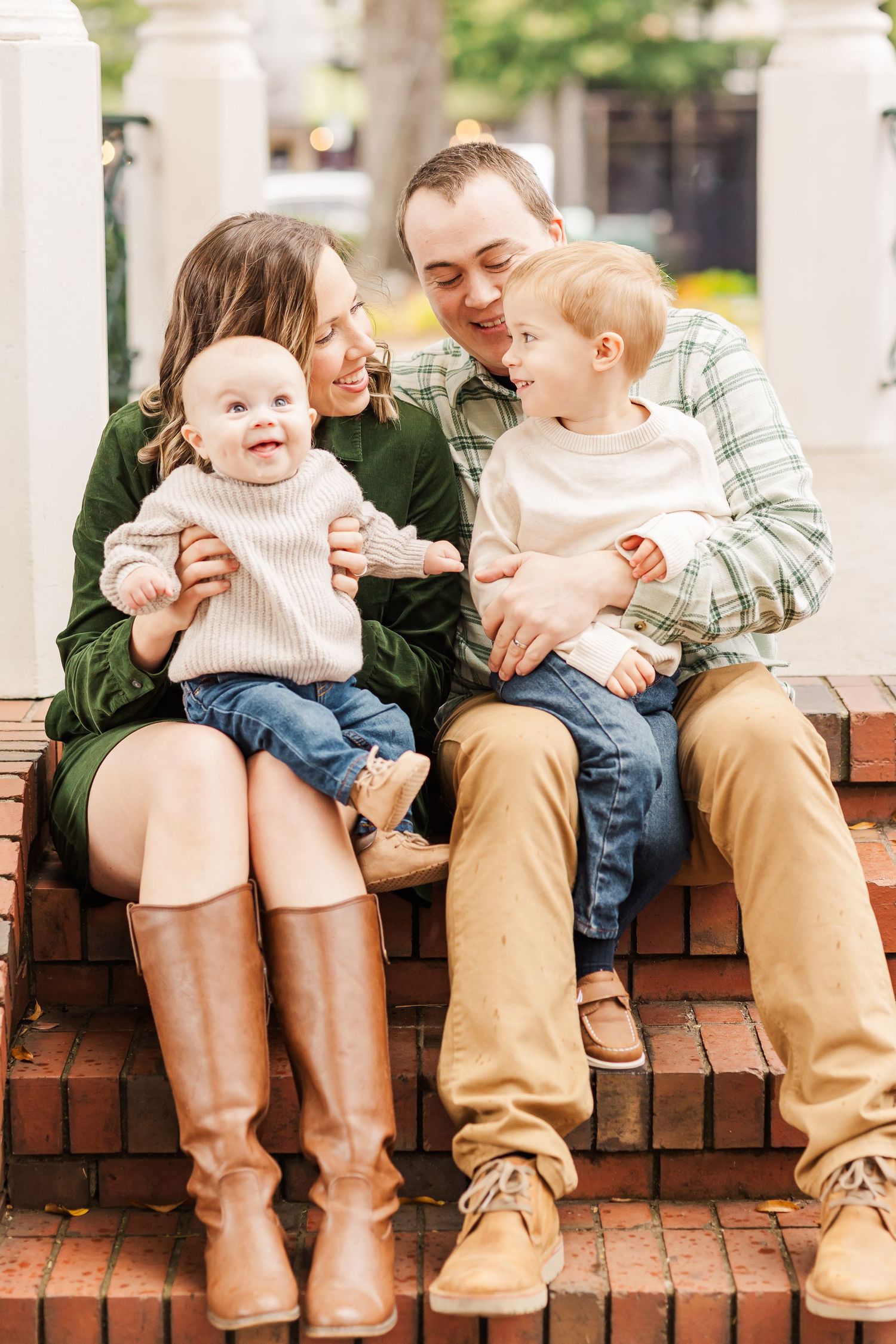 A happy family of four sits on brick steps, parents smiling at two young toddlers in a warm outdoor portrait.