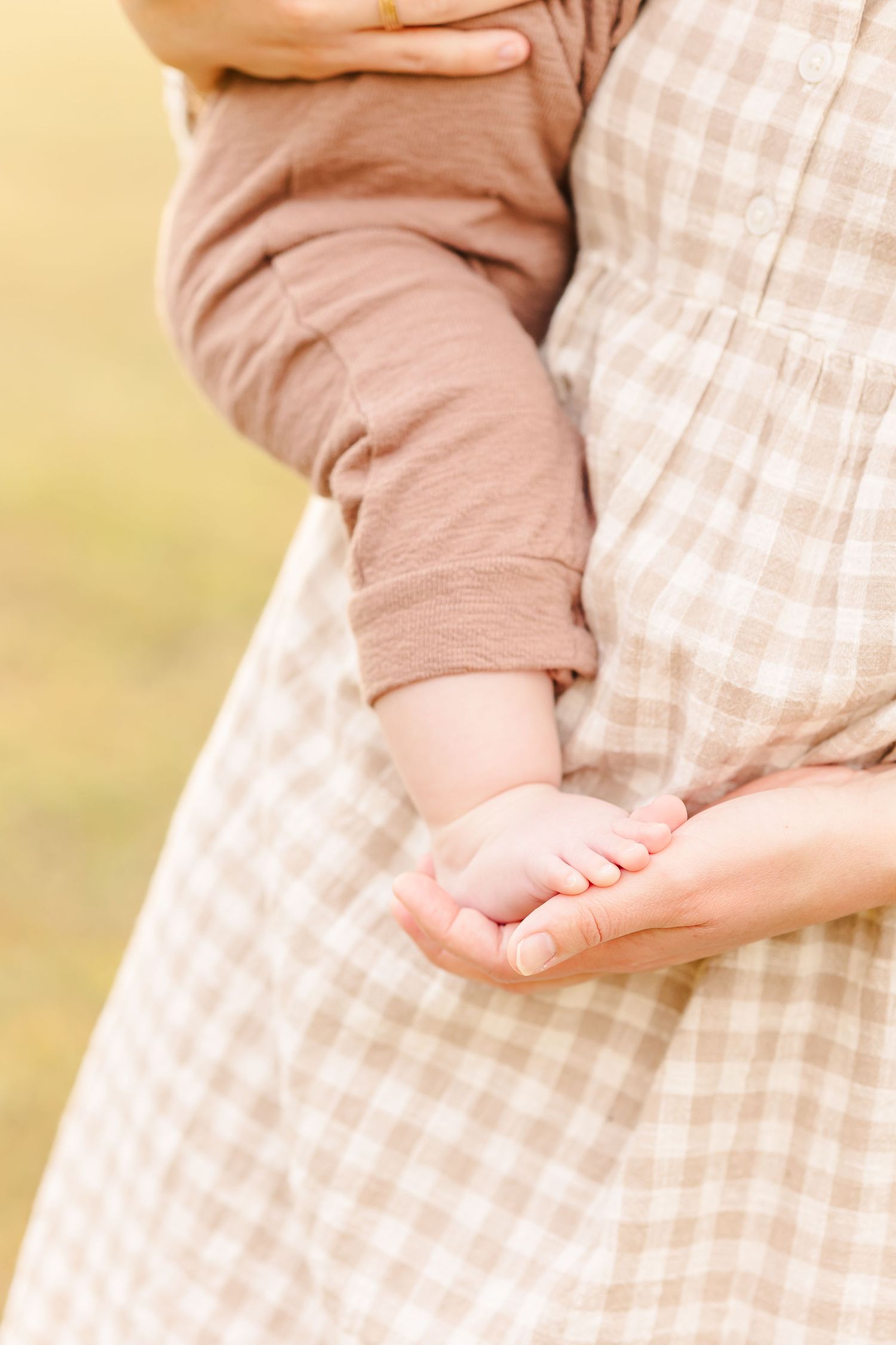 A mother in a plaid dress tenderly holds her baby's tiny feet in her hands outdoors.