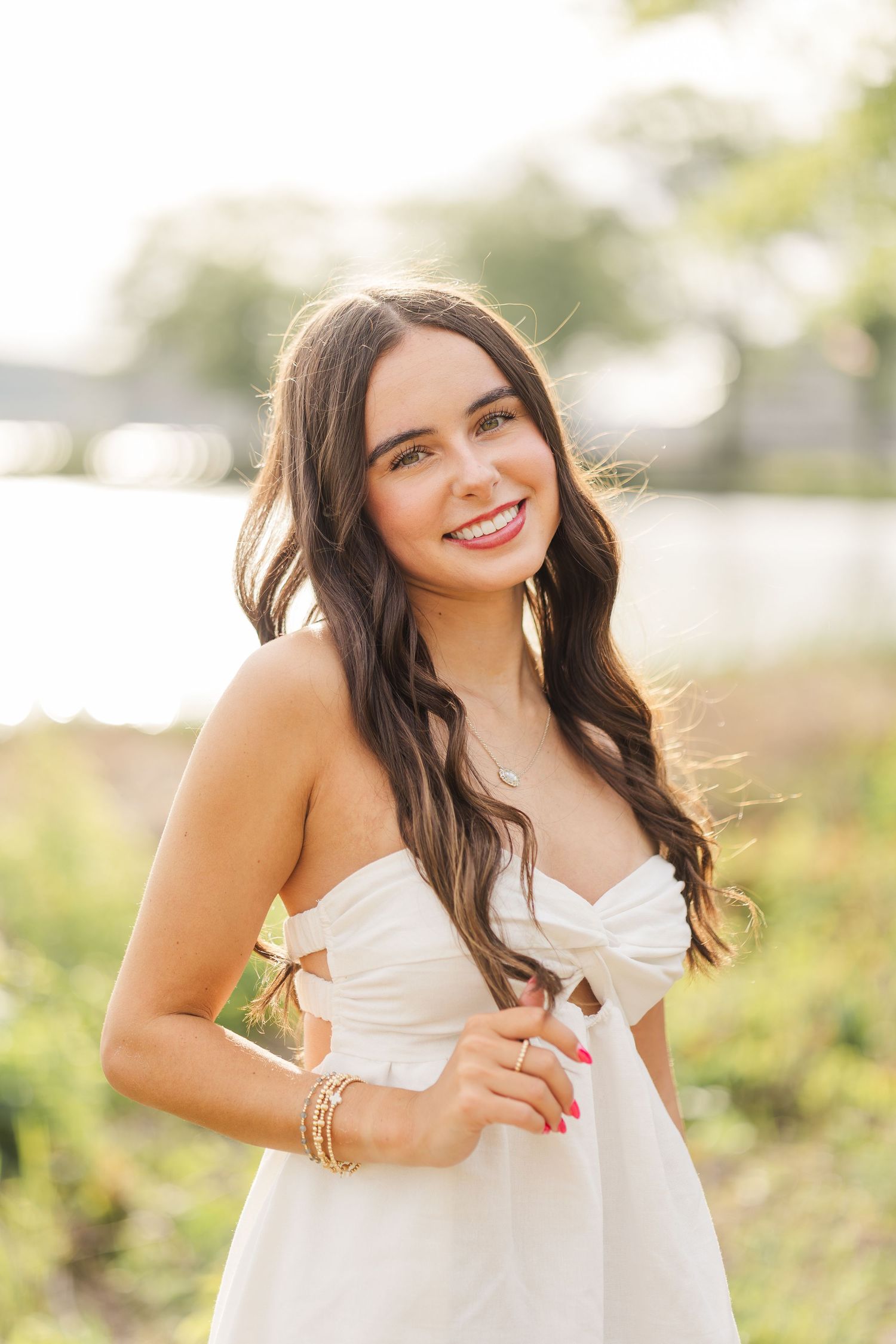 Young woman in white strapless dress smiling outdoors near water with long dark wavy hair and gold bracelets.