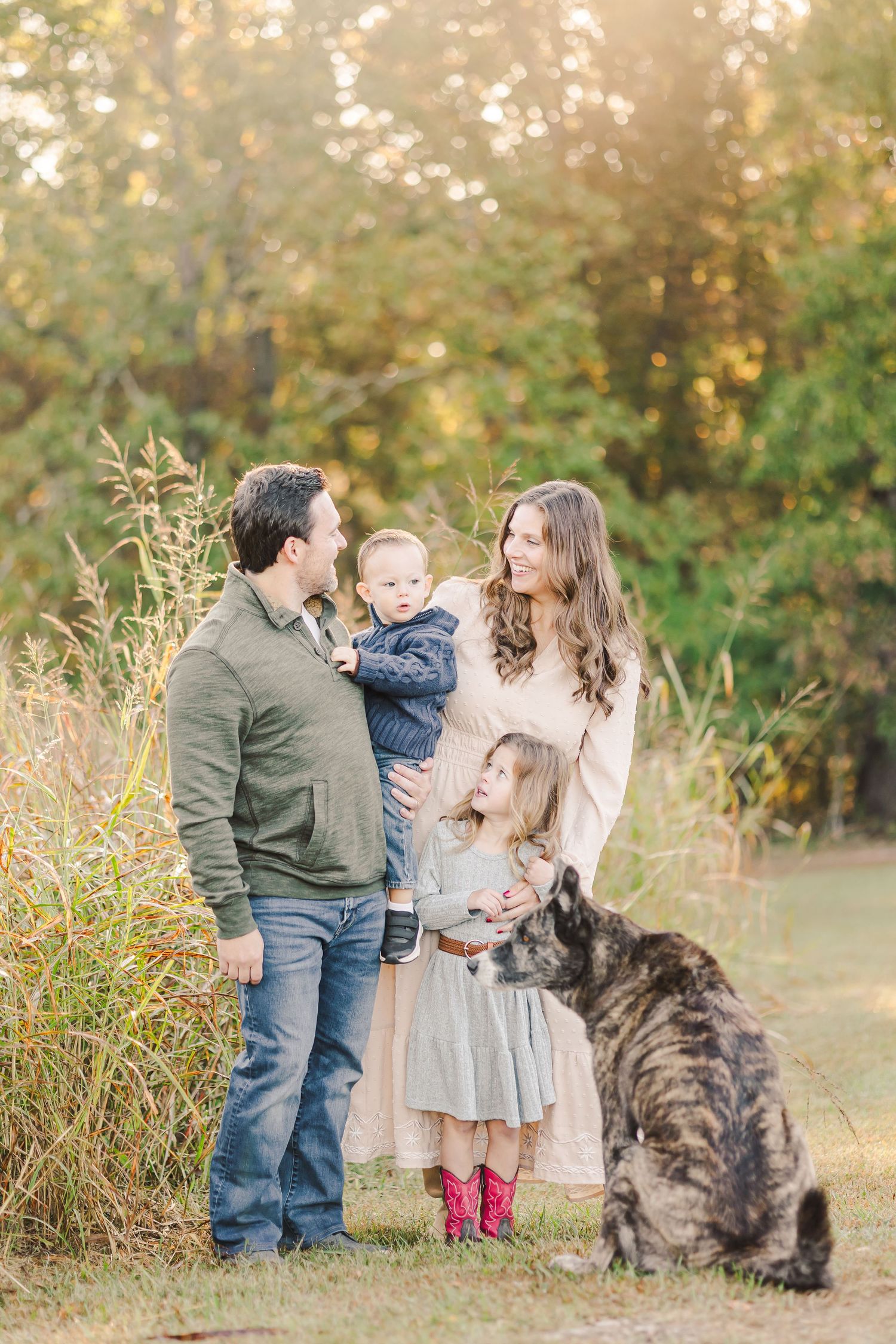 Family with two children and large dog standing in autumn field at golden hour.