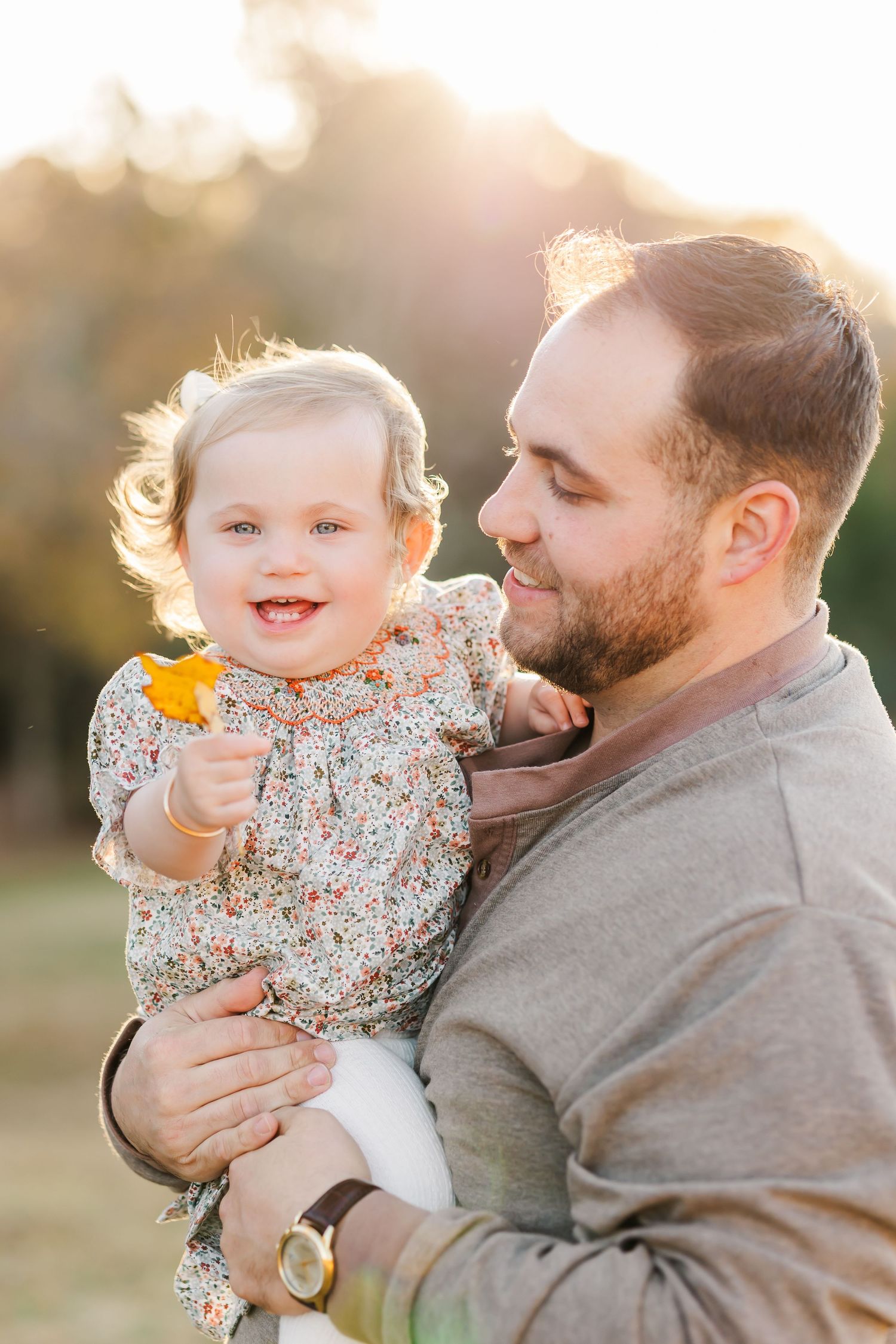 A bearded father holds his smiling baby girl outdoors in golden sunlight, as she clutches a yellow leaf.