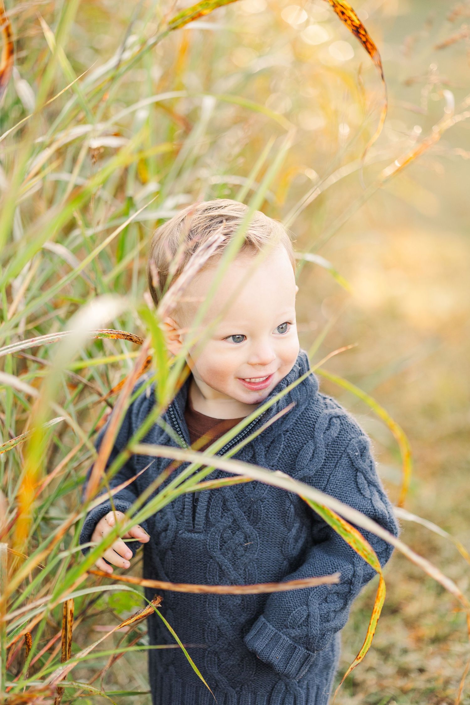 Smiling toddler boy in a navy knit sweater peeks through tall autumn grasses outdoors.