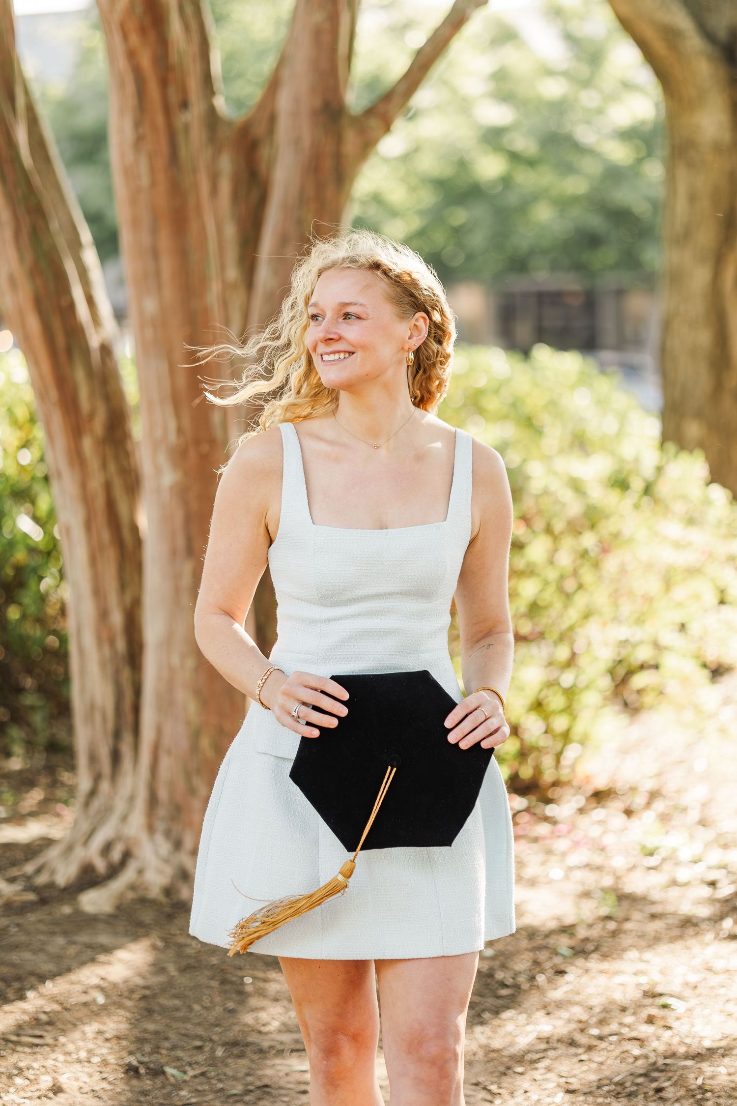 Smiling blonde woman in white dress holding graduation cap outdoors among trees on a sunny day.