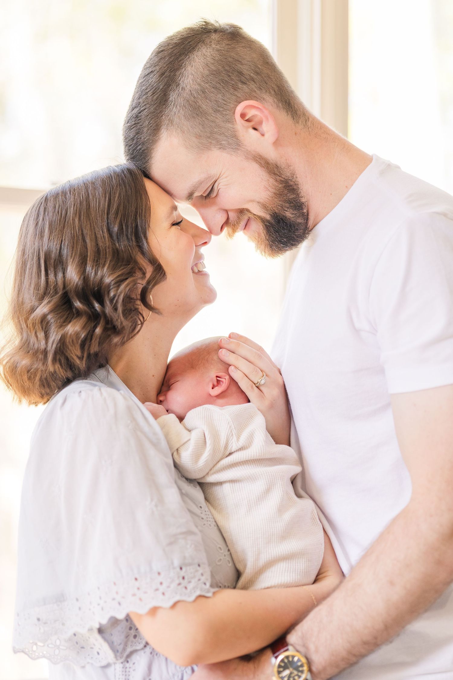 Smiling new parents touch foreheads while tenderly cradling their newborn baby indoors.