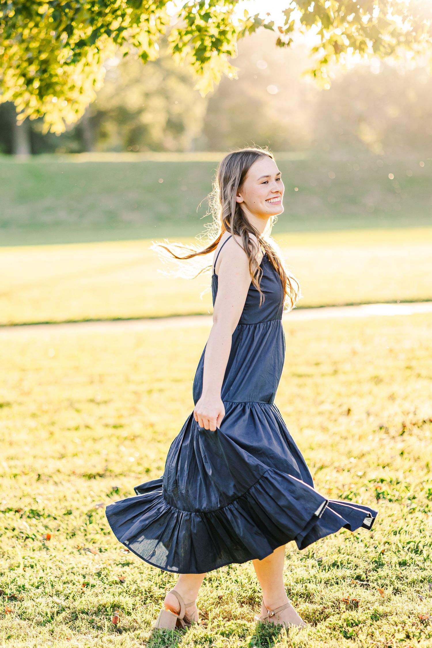 Young woman in navy blue dress twirling joyfully in a sunlit park during golden hour.