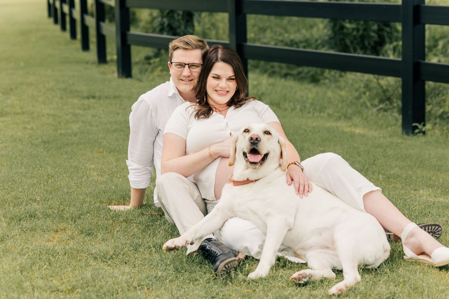 Couple in white outfits sitting on green grass with a white Labrador dog near a black fence outdoors.