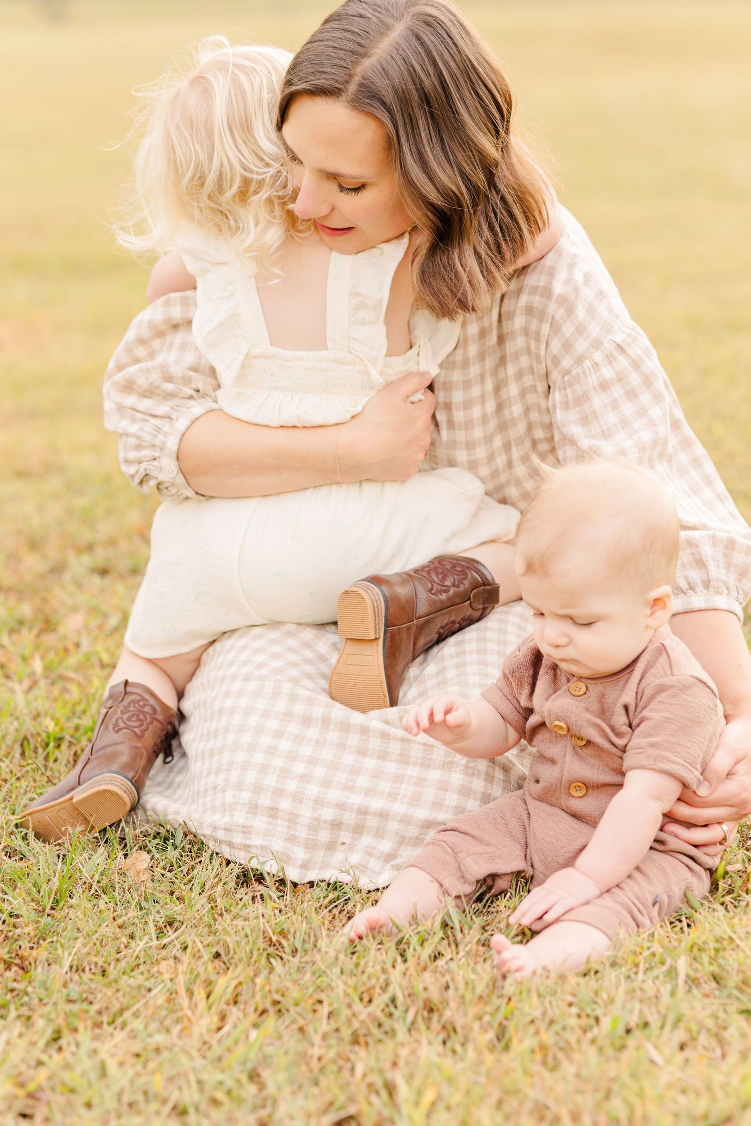 Mother sits on grass hugging her toddler while her baby boy explores during an outdoor session.