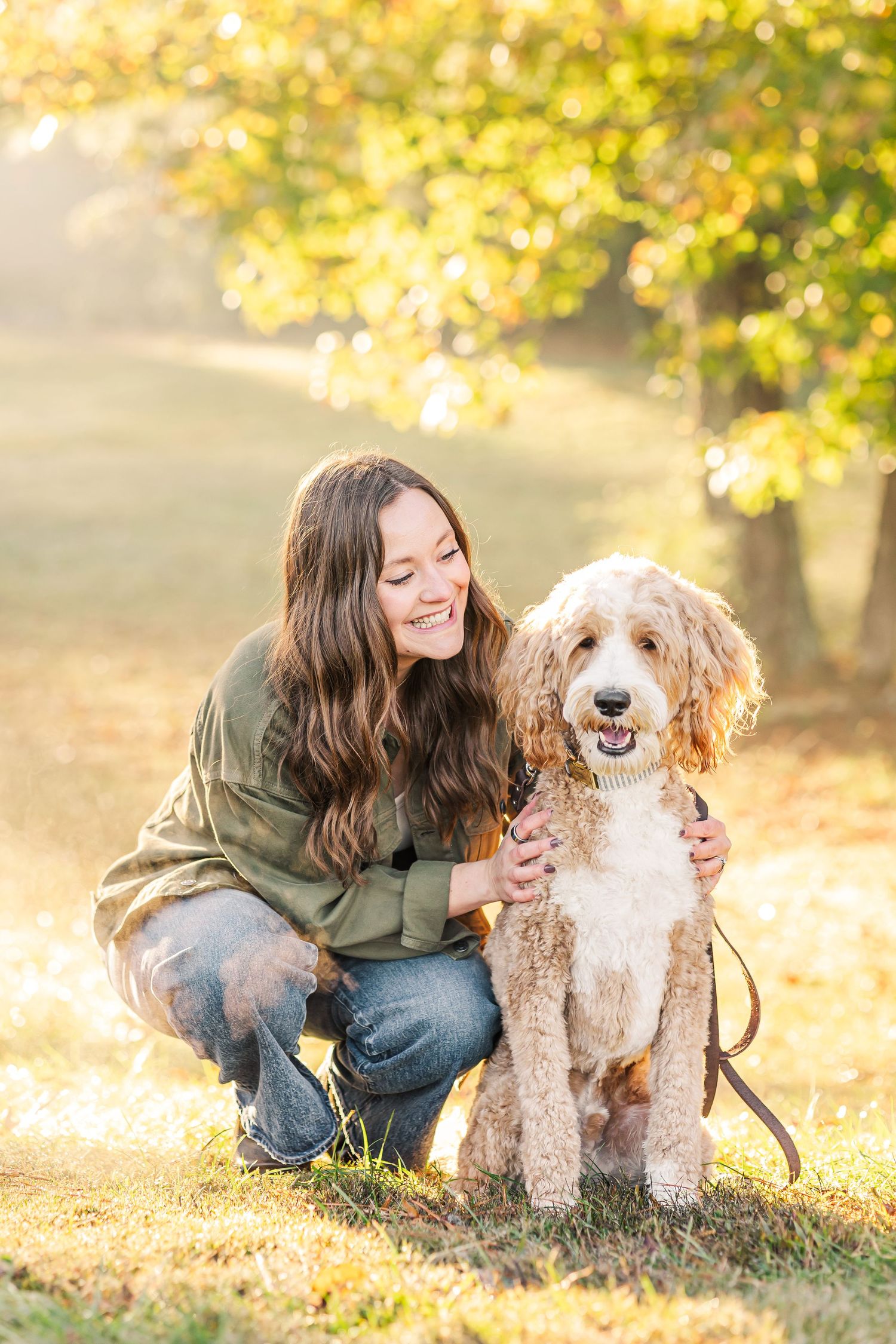 Smiling young woman crouching beside fluffy goldendoodle in golden autumn park.