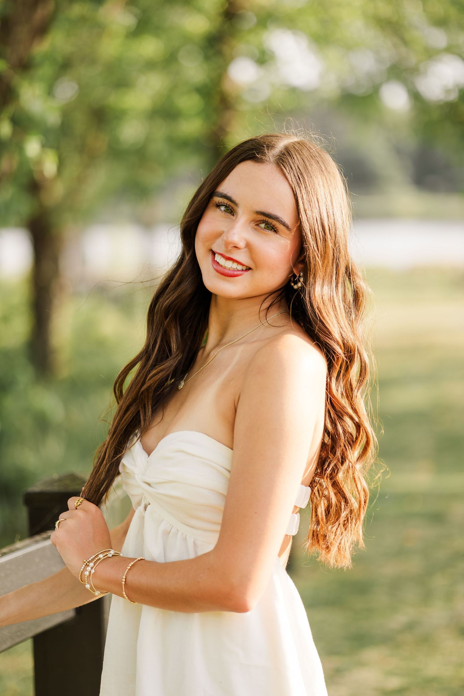 Young woman in white strapless dress with long wavy hair smiling outdoors in a sunny park setting.