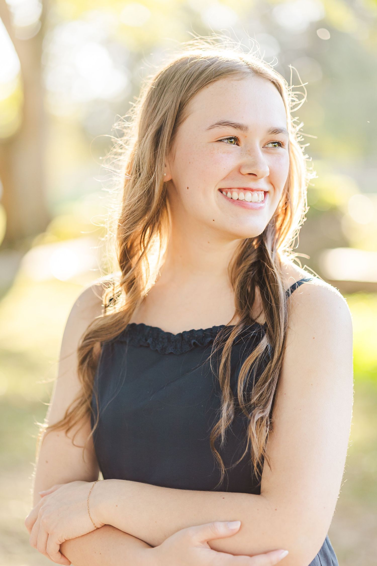 Smiling teenage girl with long hair poses outdoors during a sunny senior portrait session.
