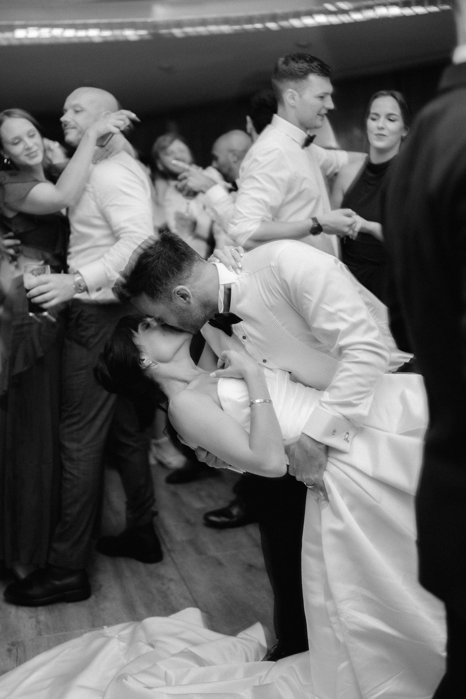 Black and white photo of a couple sharing a romantic dip kiss on the wedding dance floor surrounded by guests.