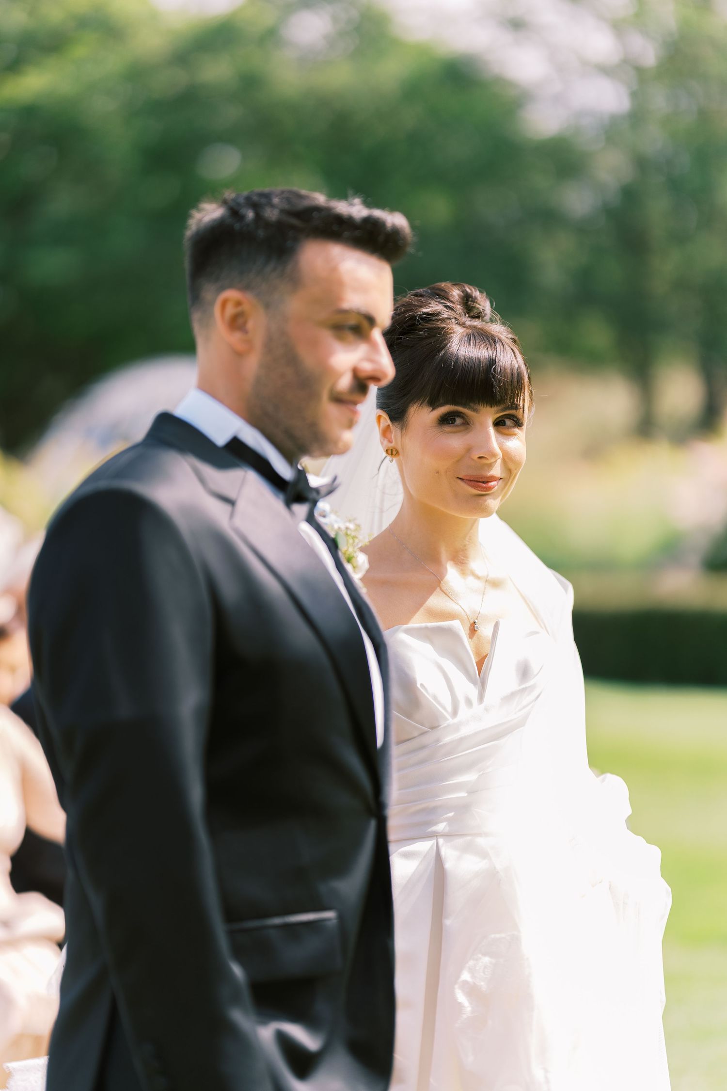 Bride in white gown and groom in black tuxedo standing together outdoors at a wedding ceremony.