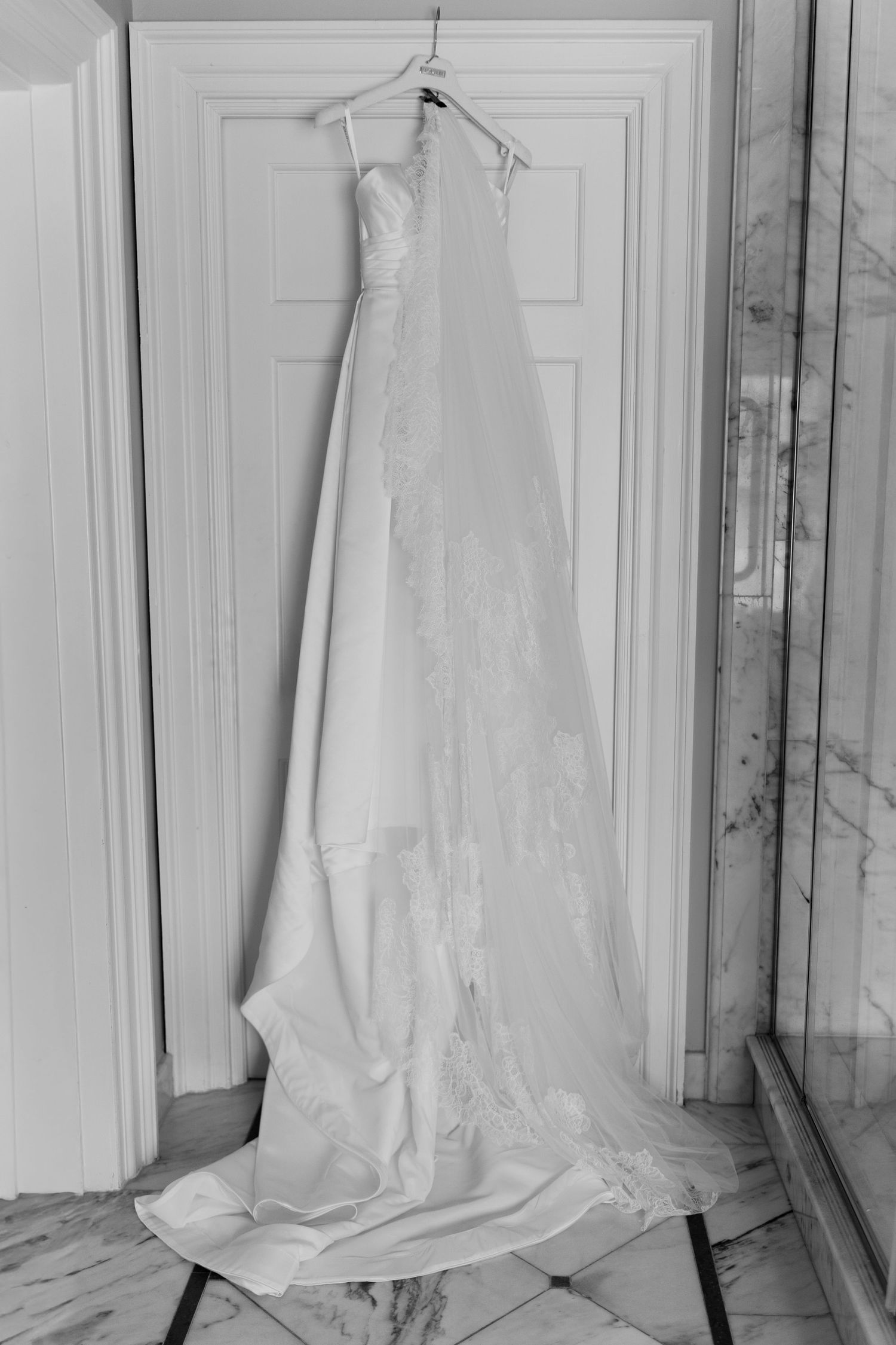 Elegant white wedding dress with lace detail and long train hanging on a door hanger in black and white photo.