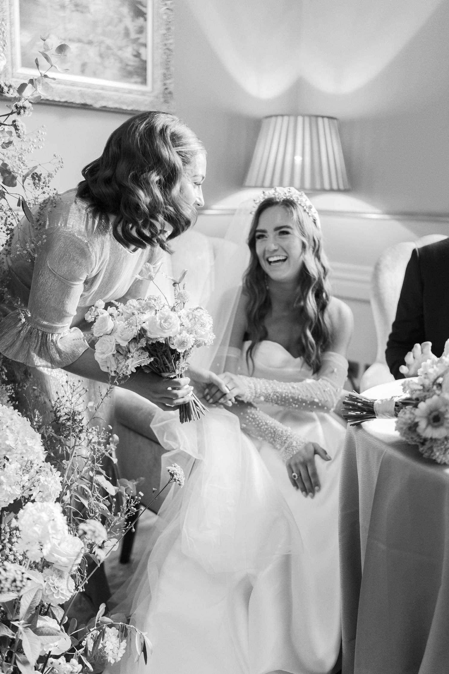 Black and white photo of a laughing bride in a strapless gown and veil, sharing a joyful moment with a bridesmaid holding flowers.