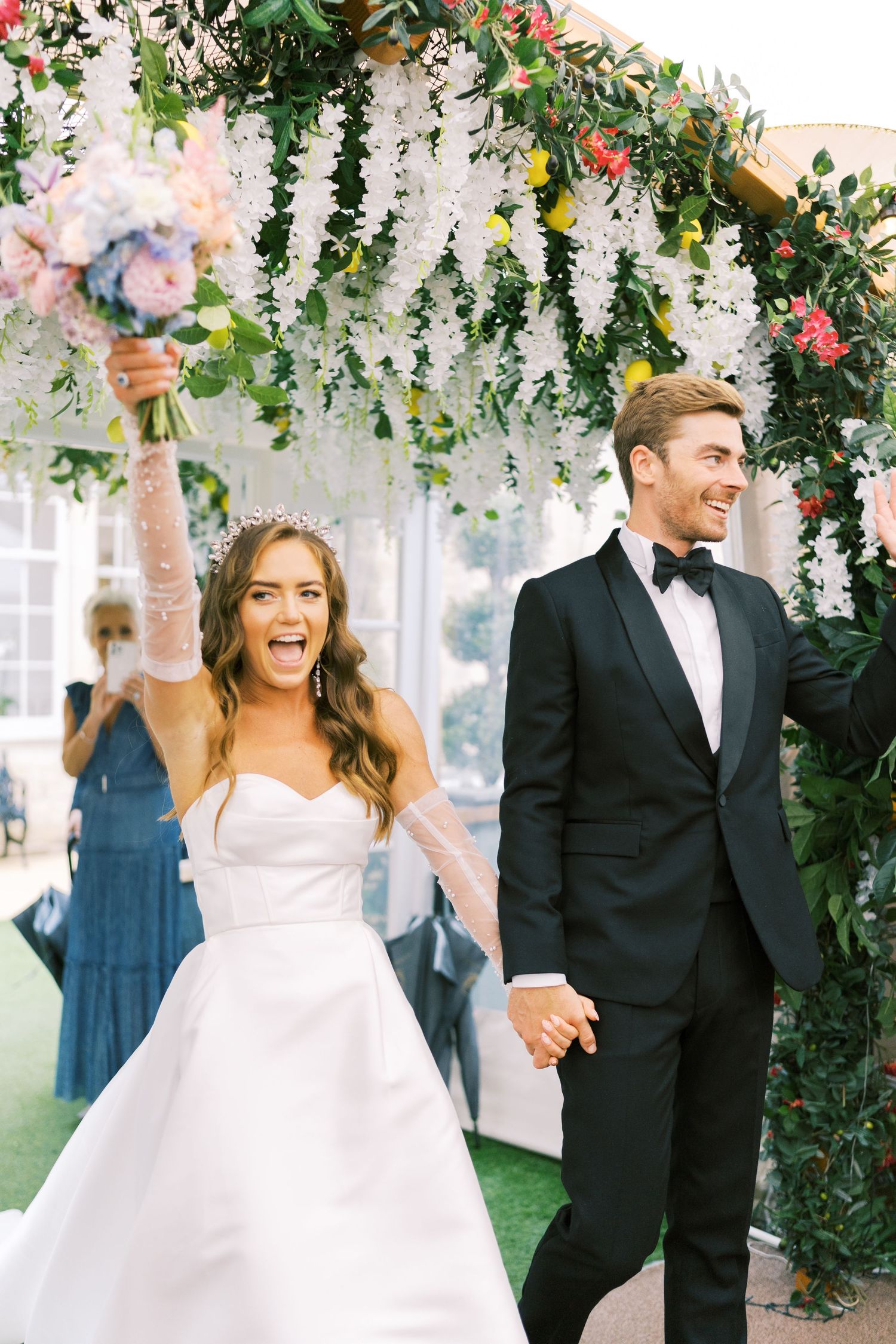 Joyful bride and groom celebrate under floral arch, bride raising bouquet, groom in black tuxedo, both smiling.