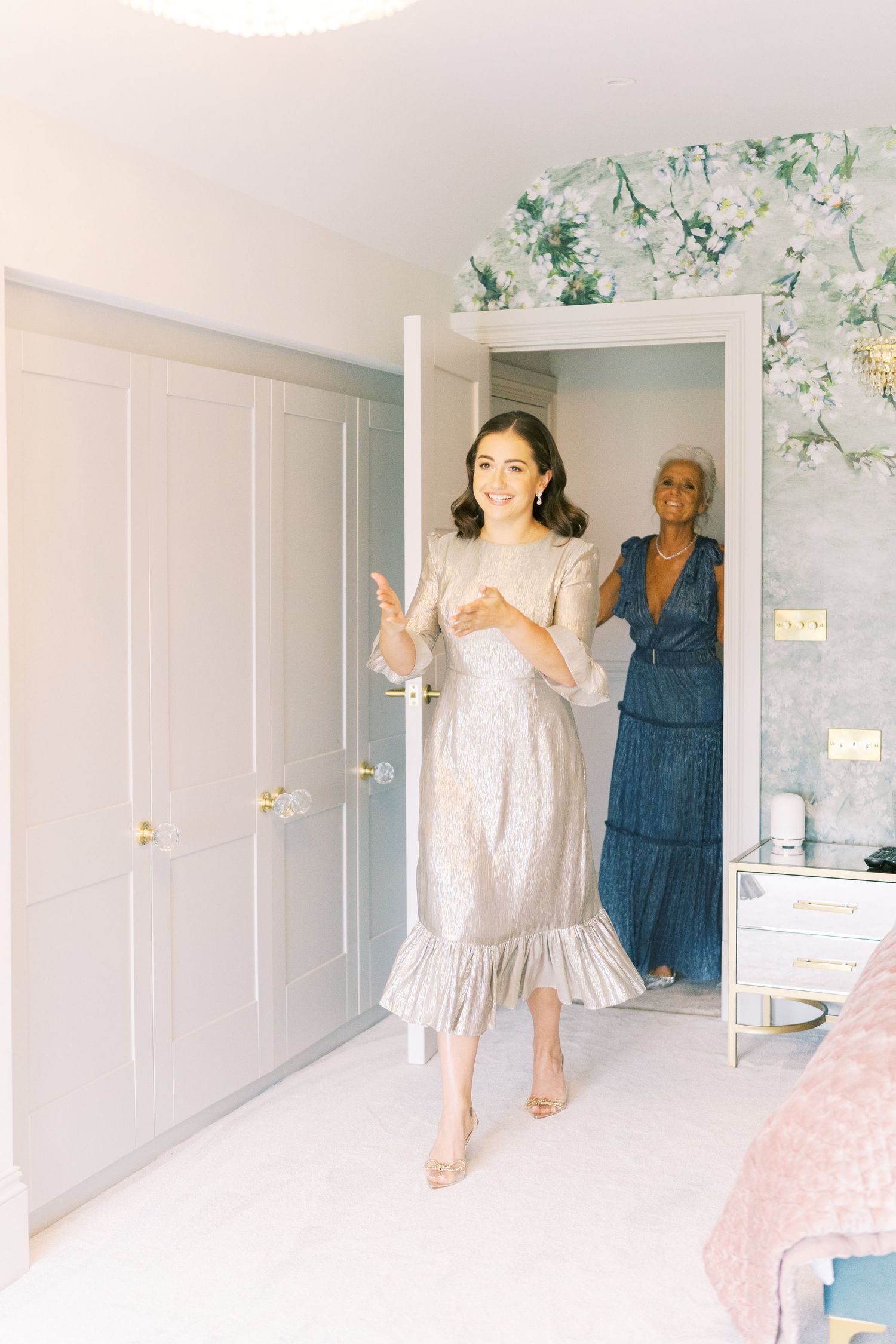 Bride in silver midi dress walks through doorway smiling, with woman in blue dress visible behind her in elegant room.
