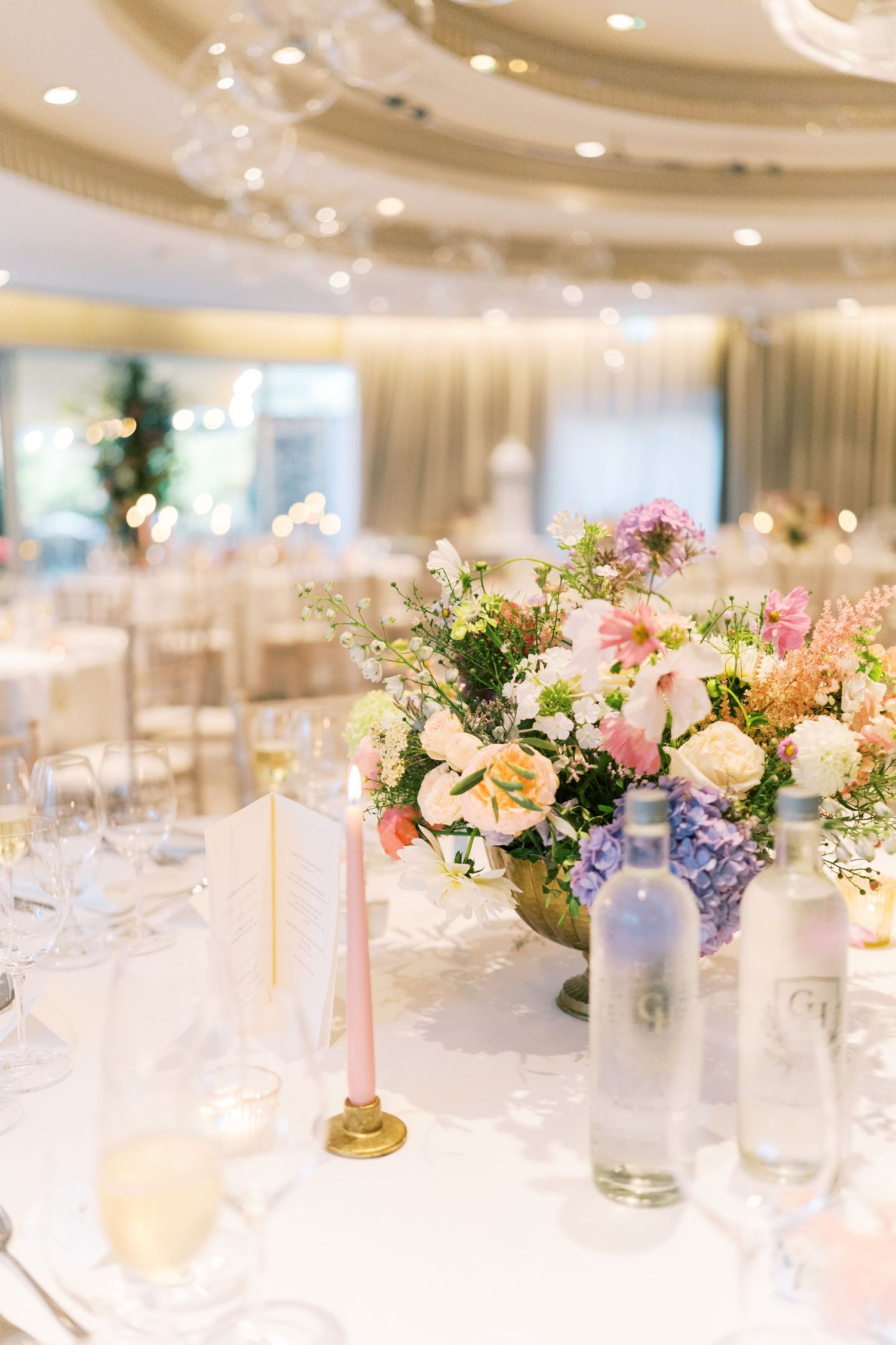 Elegant wedding reception table with pastel floral centerpiece, pink taper candle, glassware, and water bottles in a bright ballroom.