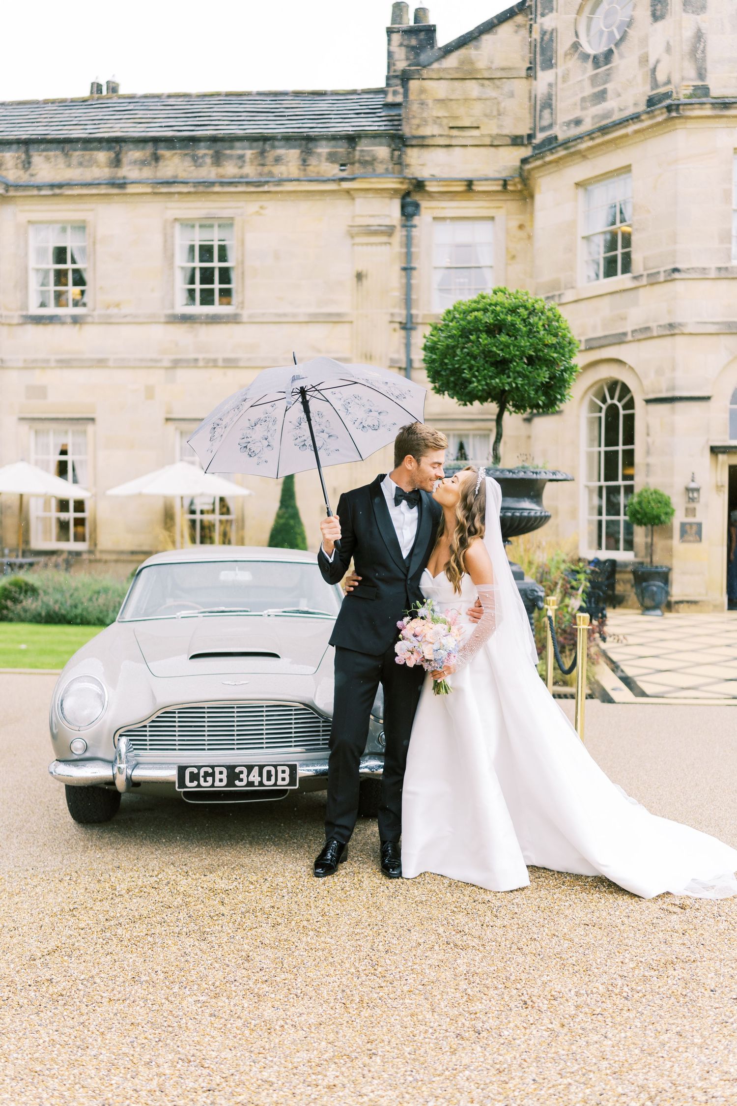 Bride and groom share a romantic moment under an umbrella beside a vintage Aston Martin outside an elegant manor house.