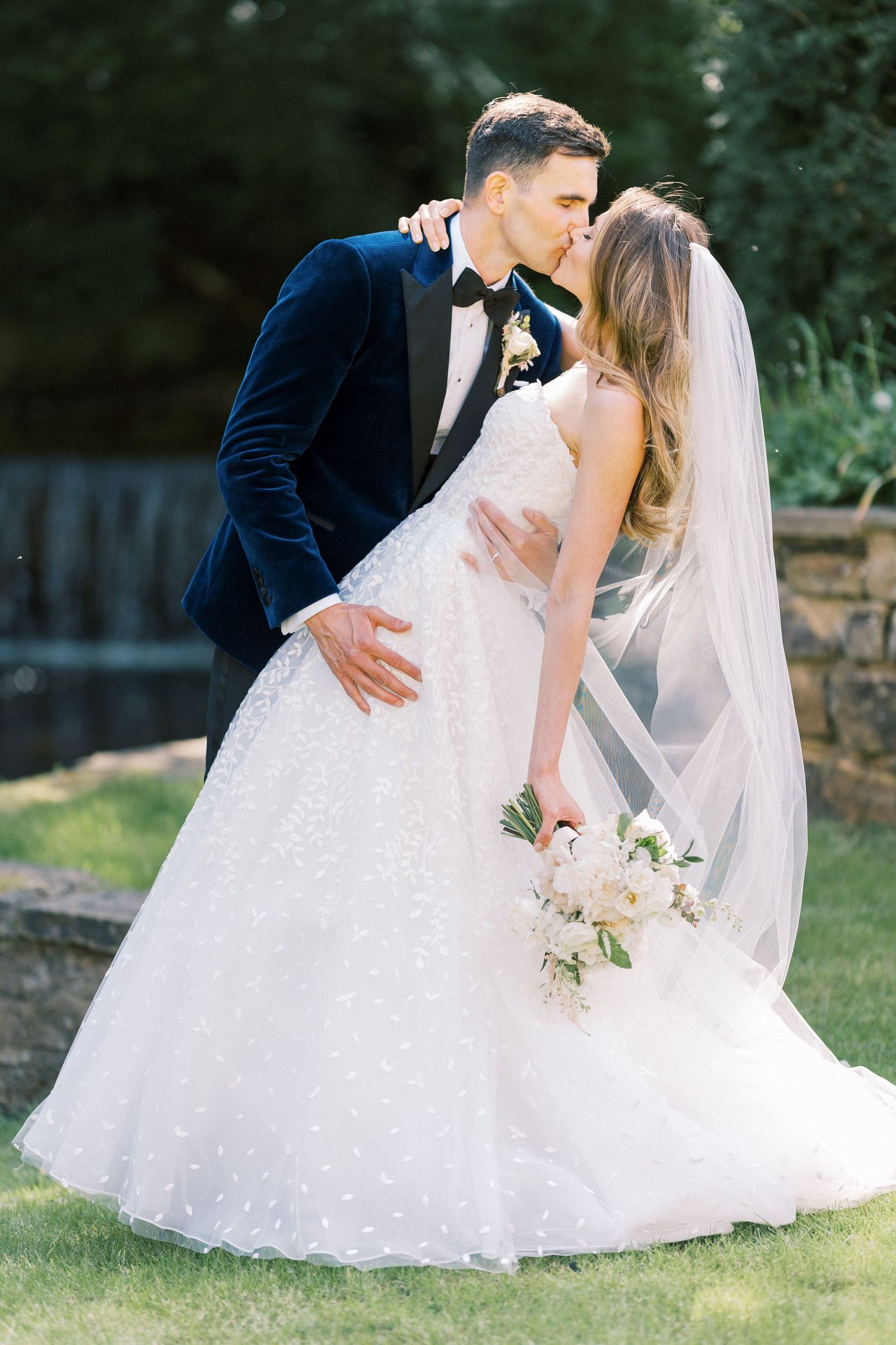 Bride and groom share a romantic kiss outdoors, she holds a white bouquet in a ballgown with veil, he wears a navy tuxedo.
