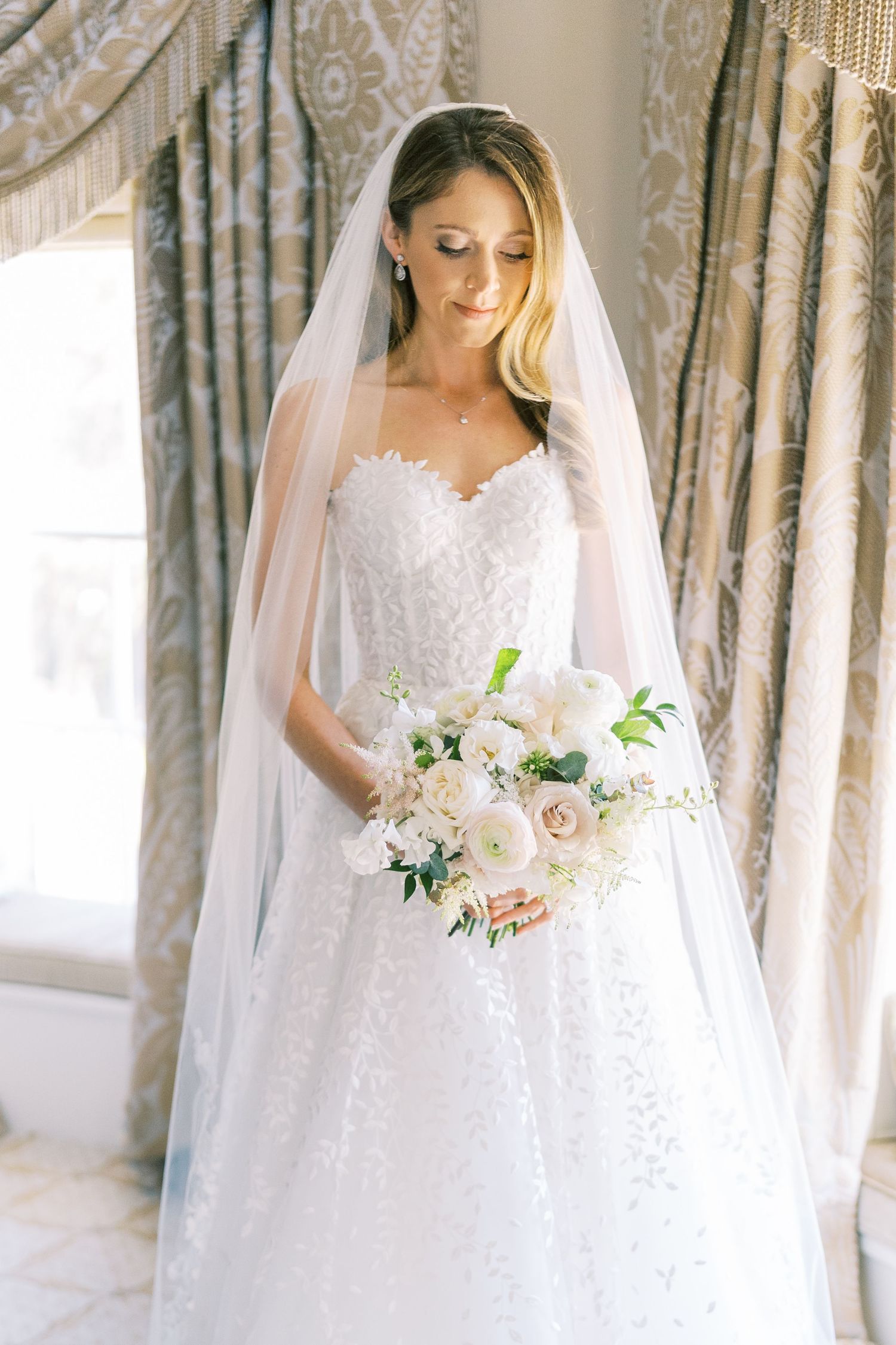 Elegant bride in strapless lace ballgown and cathedral veil holds white floral bouquet by window.