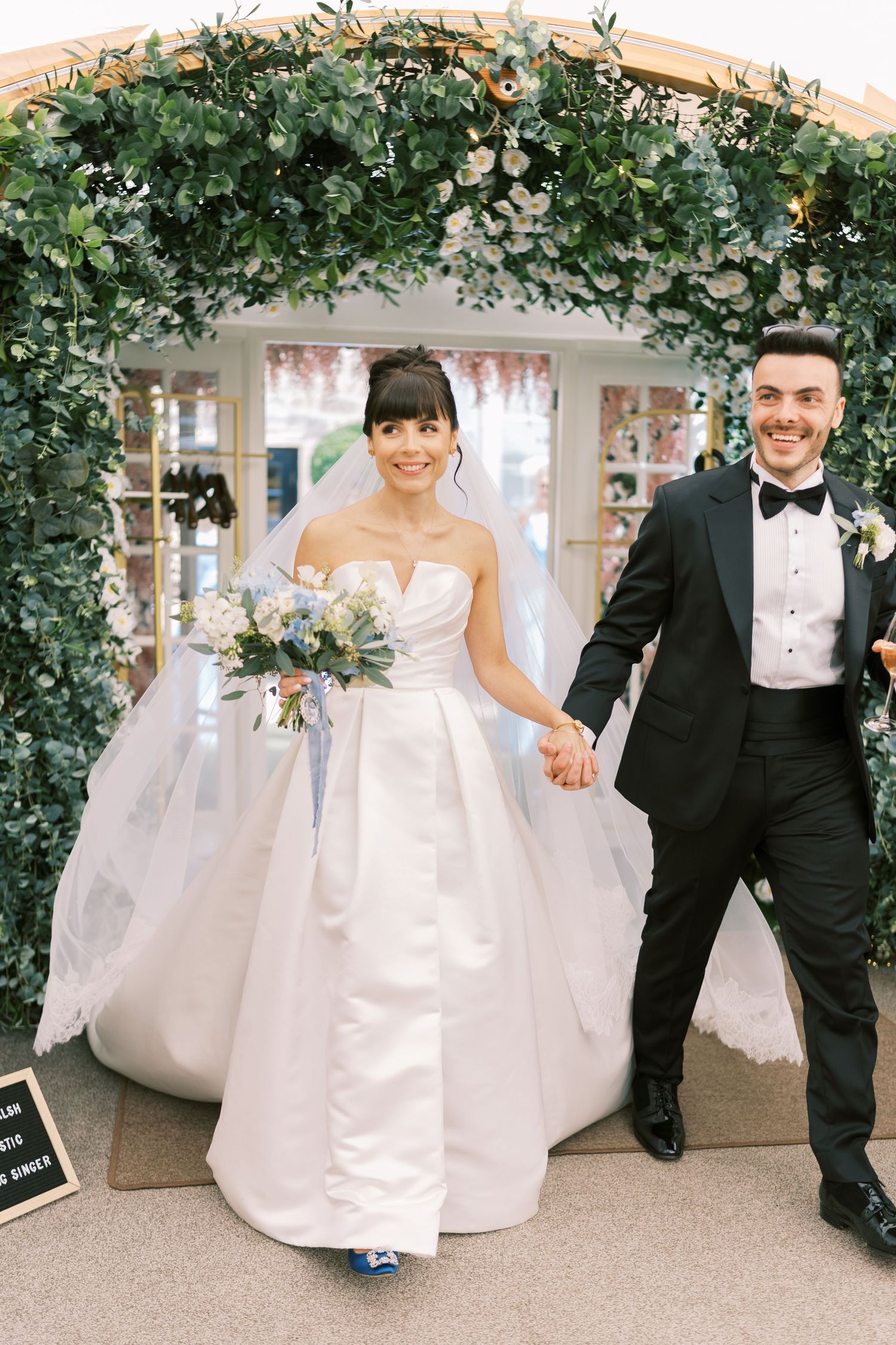 Smiling bride and groom holding hands under a lush greenery arch, bride in strapless ball gown holding blue bouquet.