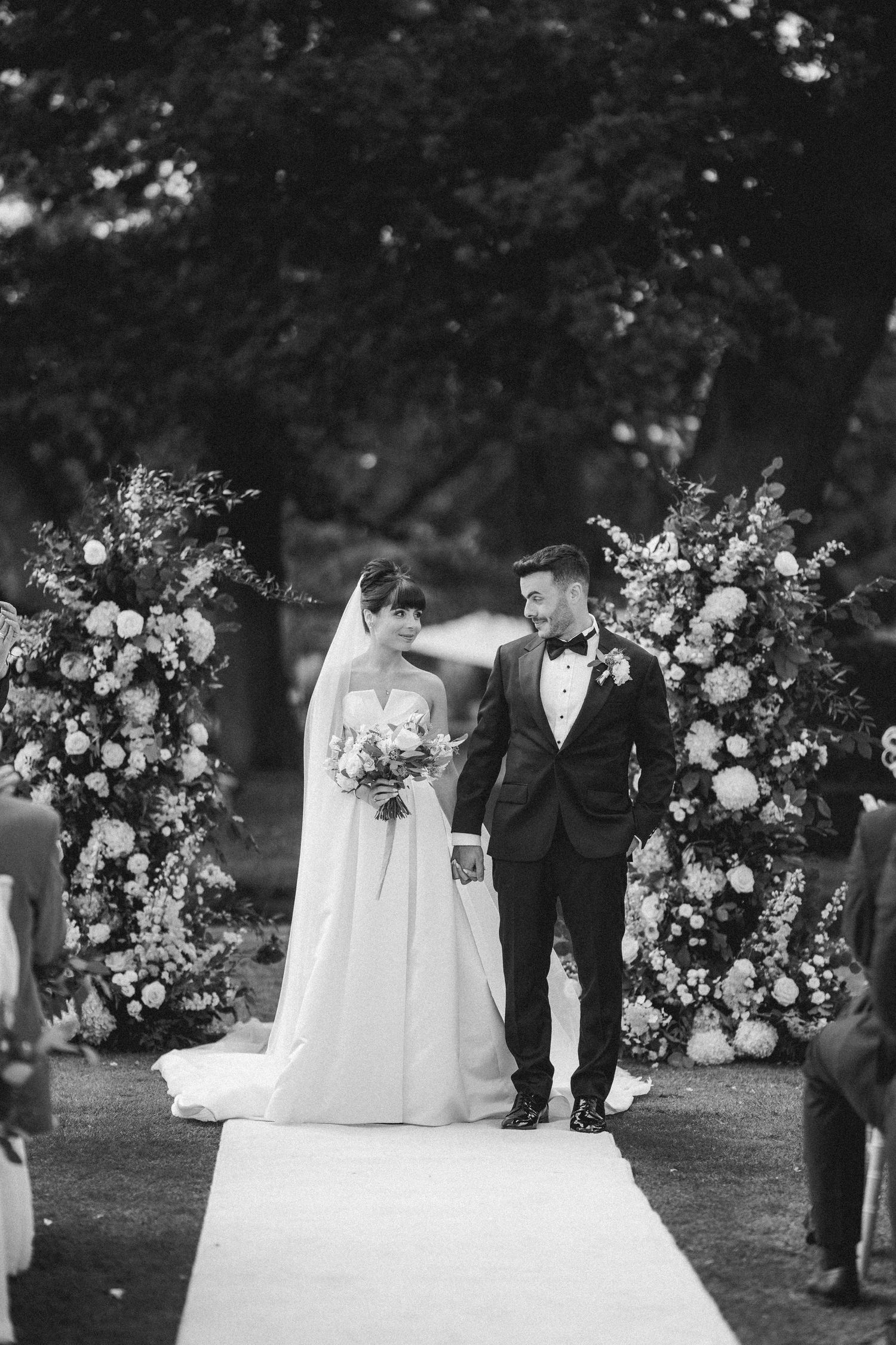 Bride and groom walk down aisle flanked by floral arrangements at outdoor wedding ceremony.