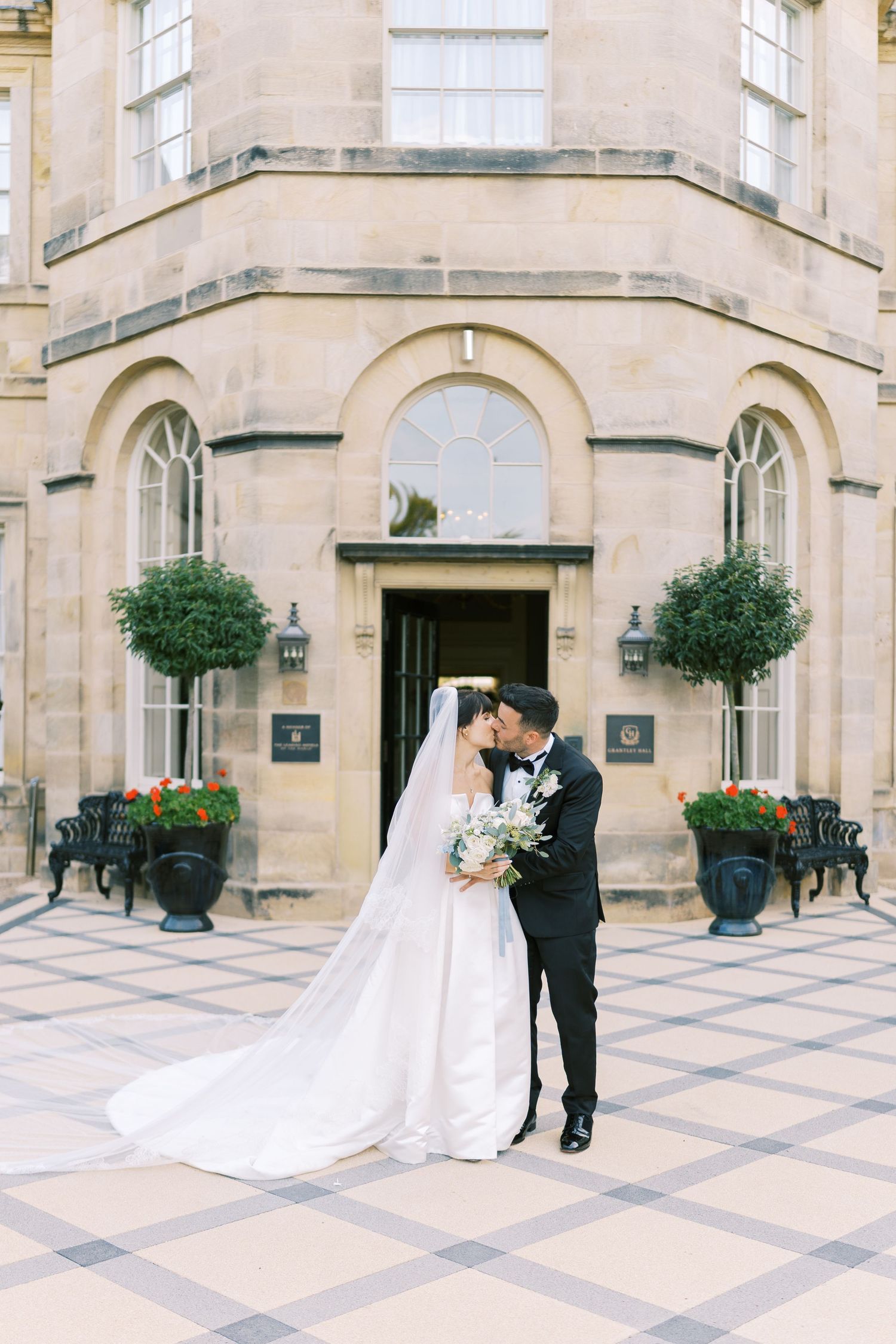 Bride and groom kissing outside a grand stone building, bride in flowing white gown with veil, groom in black tuxedo.