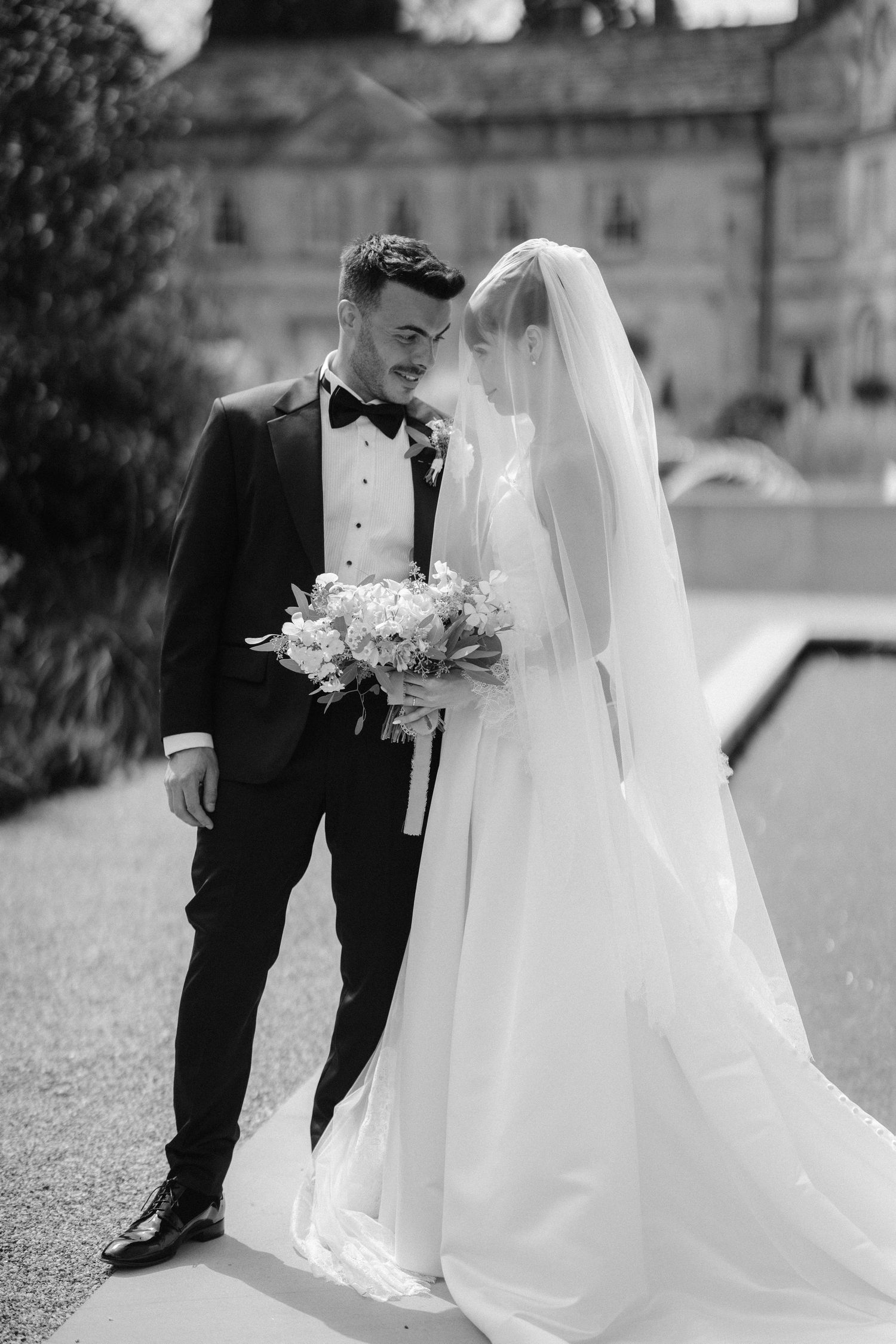 Black and white photo of bride and groom sharing intimate moment, groom in tuxedo, bride in flowing gown with veil.