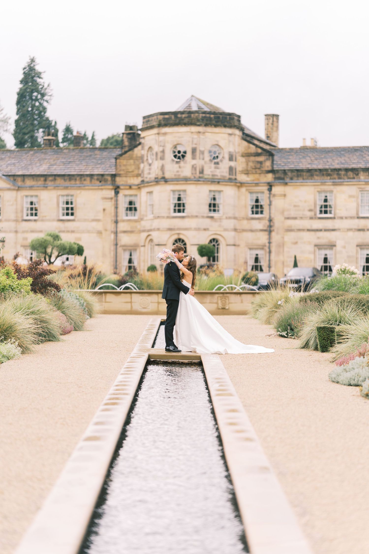 Bride and groom sharing a kiss beside a reflecting pool in formal gardens of a grand English country estate.