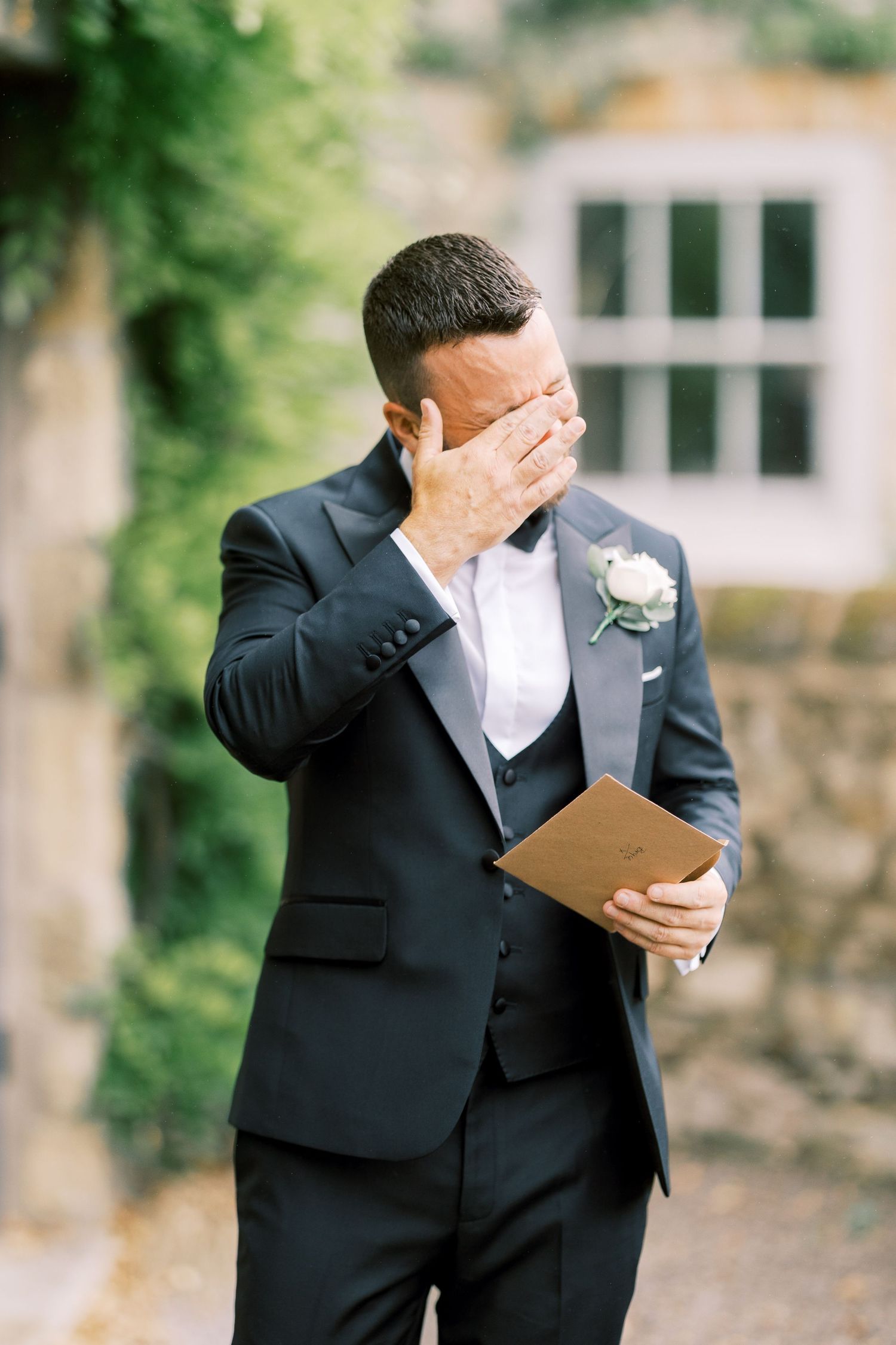 Emotional groom in navy suit wiping tears while holding wedding vows card outside stone venue with white boutonniere.