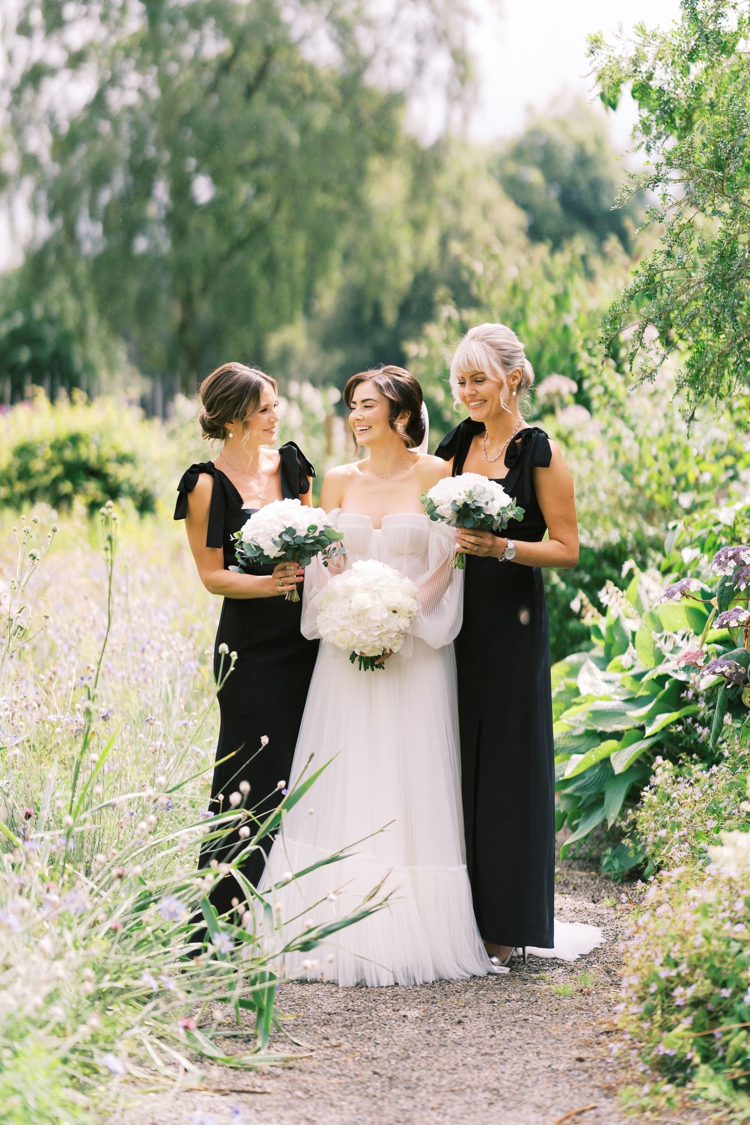 Bride in white tulle gown with two bridesmaids in black dresses holding white bouquets in a garden path.
