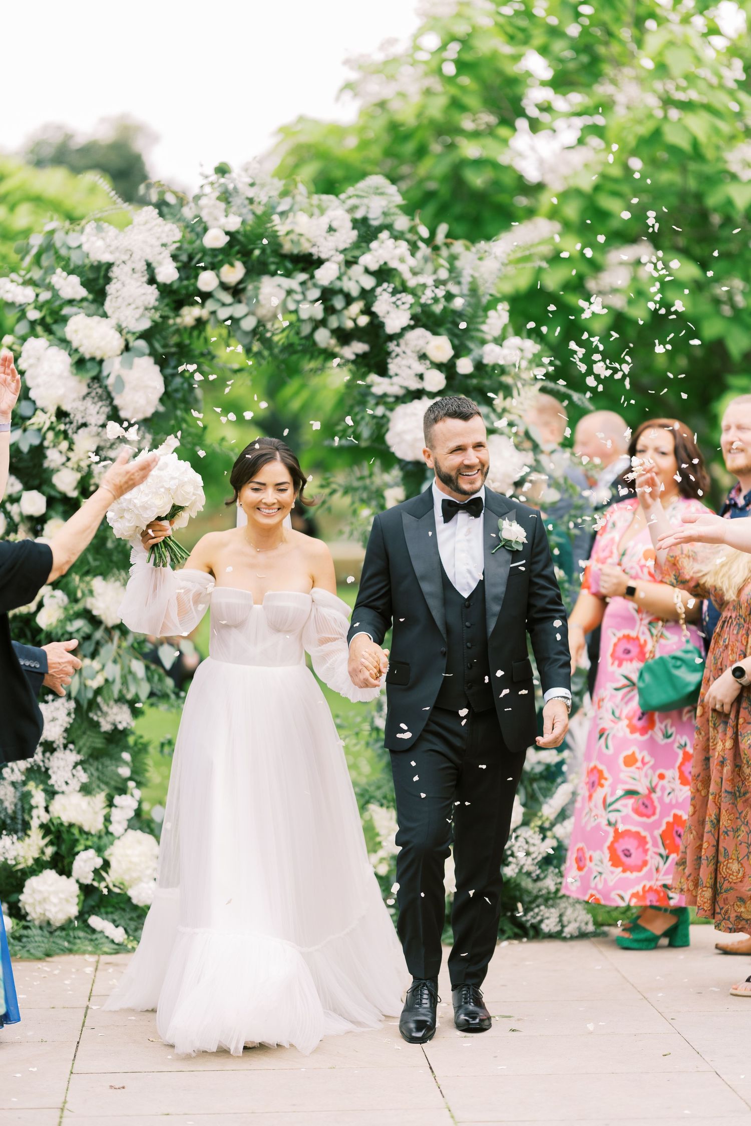 Bride and groom walk through confetti shower after wedding ceremony, smiling joyfully before floral arch backdrop.