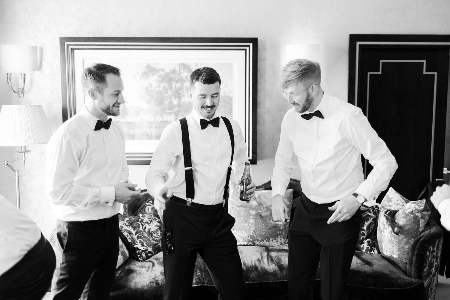 Three groomsmen in white shirts, suspenders, and bow ties laughing together in black and white photo.