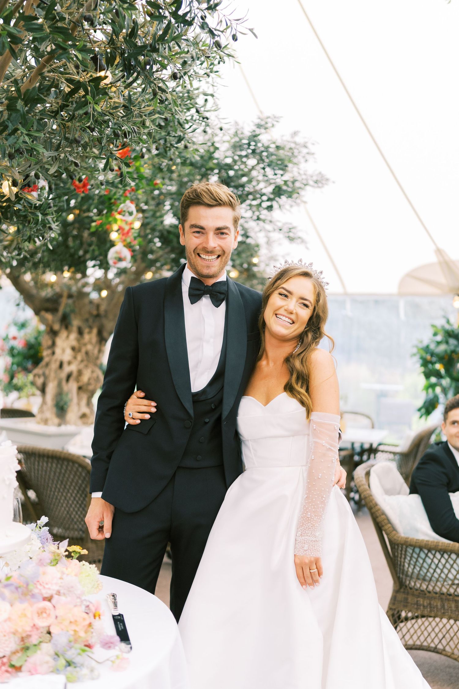 Smiling bride and groom pose together under an olive tree, she in a strapless gown, he in a black tuxedo.