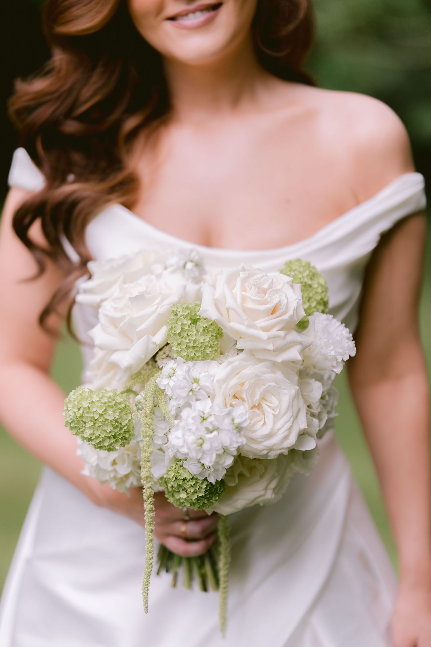 Bride in off-shoulder white gown holding elegant bouquet of white roses and green hydrangeas.