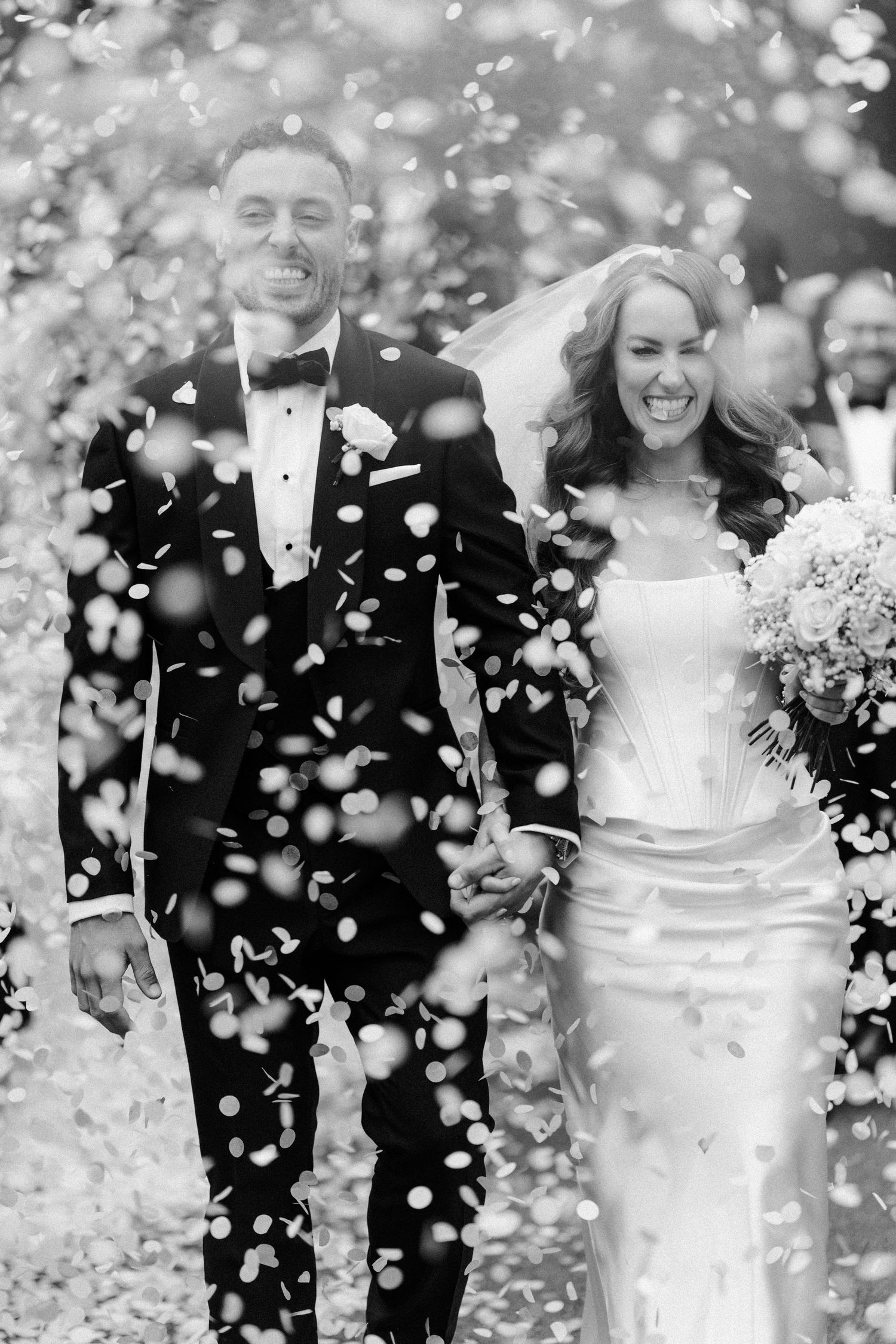 Black and white photo of a joyful bride and groom walking through confetti shower on their wedding day.