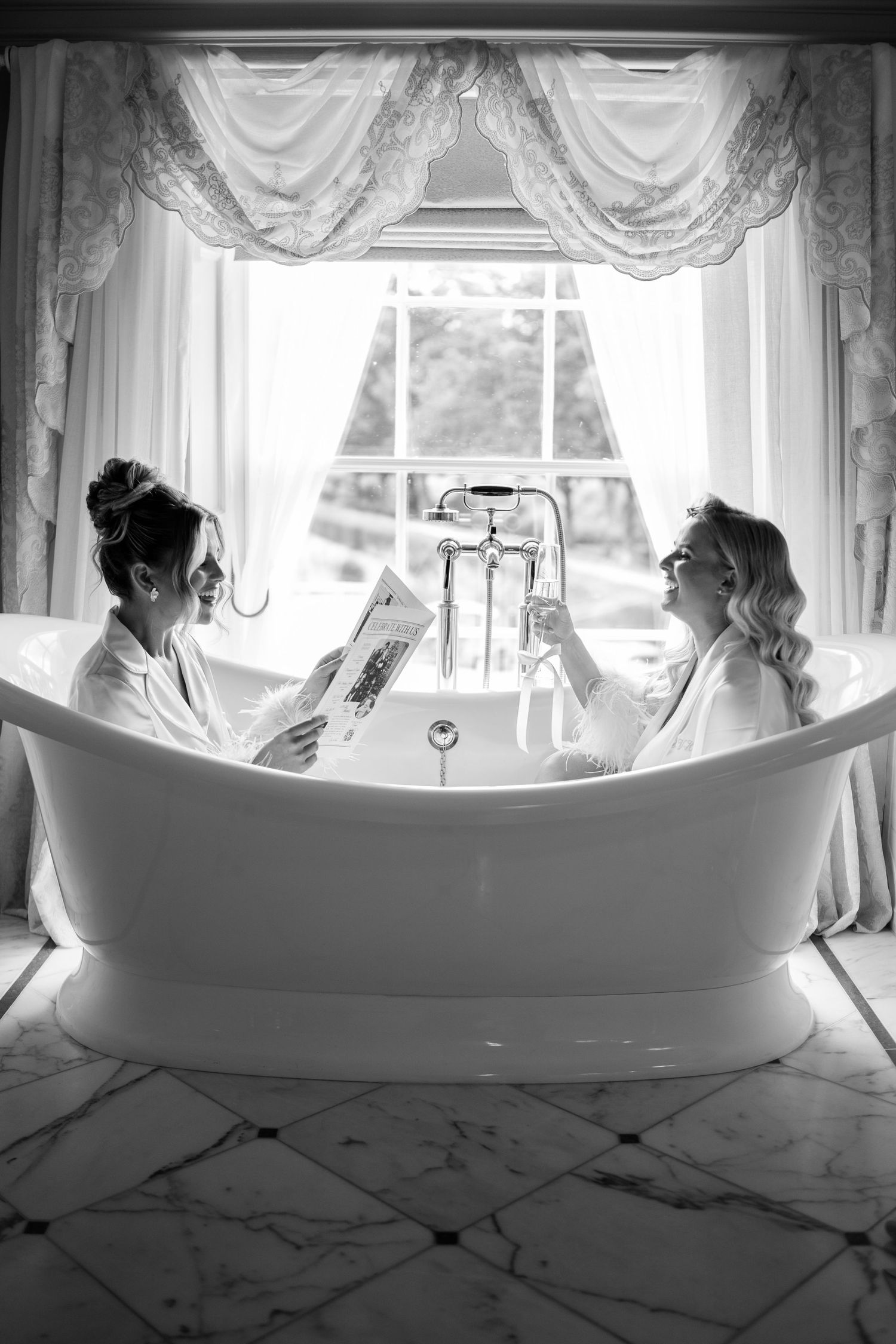 Two women in robes relaxing in a freestanding bathtub, reading newspapers in an elegant bathroom, black and white photo.
