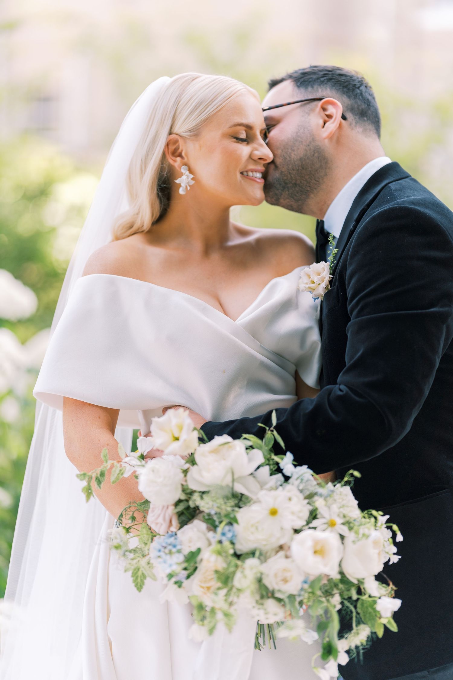 Groom tenderly kisses bride's cheek as she smiles holding a white and blue bouquet.