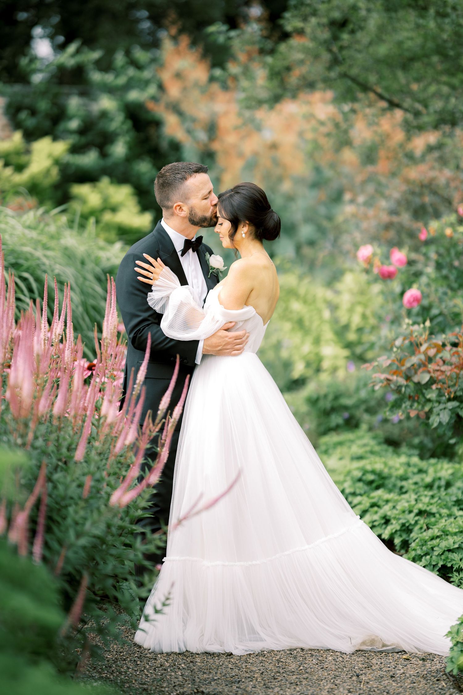 Romantic couple shares a kiss in a lush garden, bride in flowing white gown with sheer sleeves.