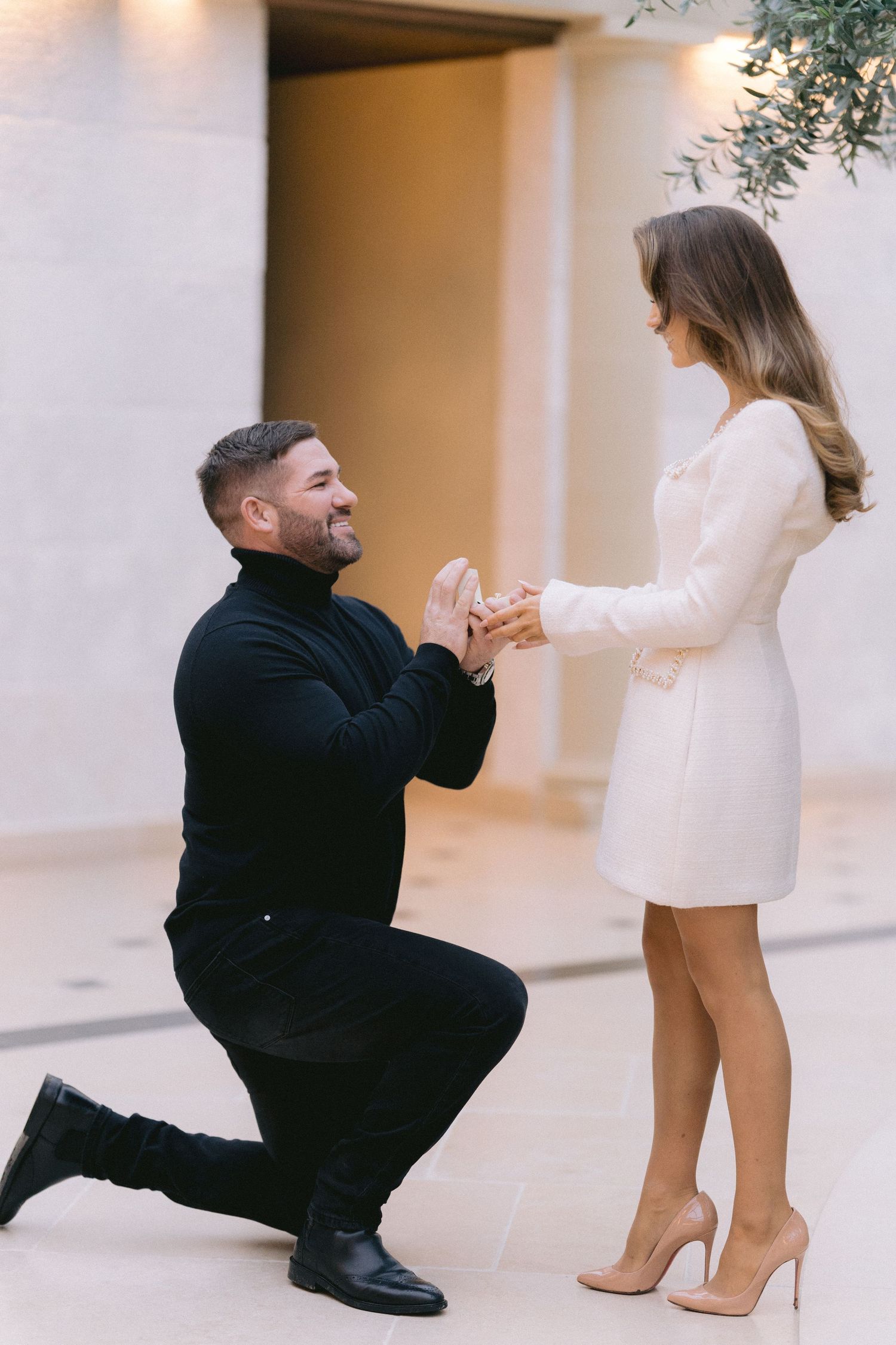 Smiling man kneeling on one knee holding womans hands during romantic marriage proposal outdoors.