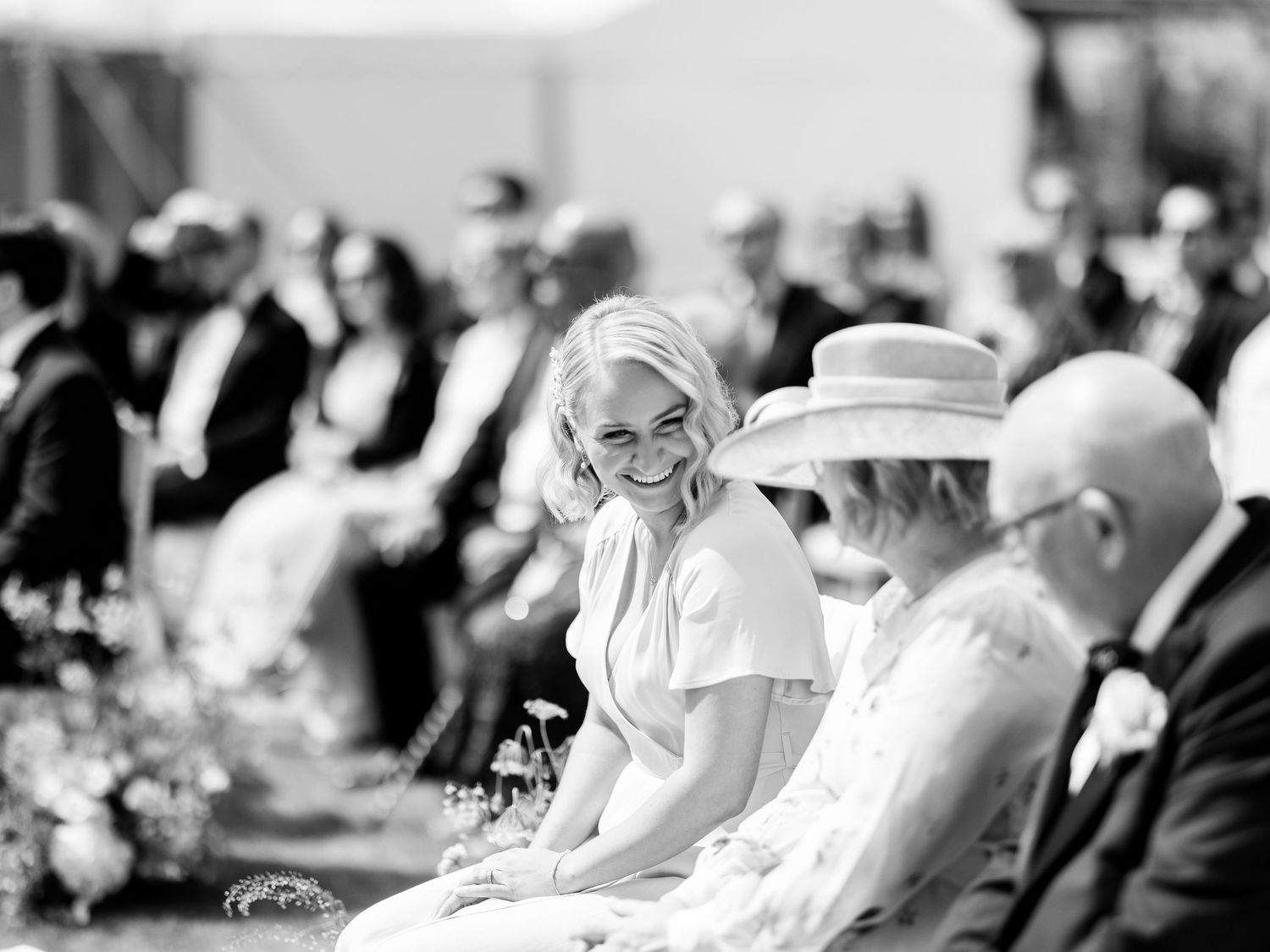 Smiling blonde bride seated among wedding guests at outdoor ceremony in black and white photo.