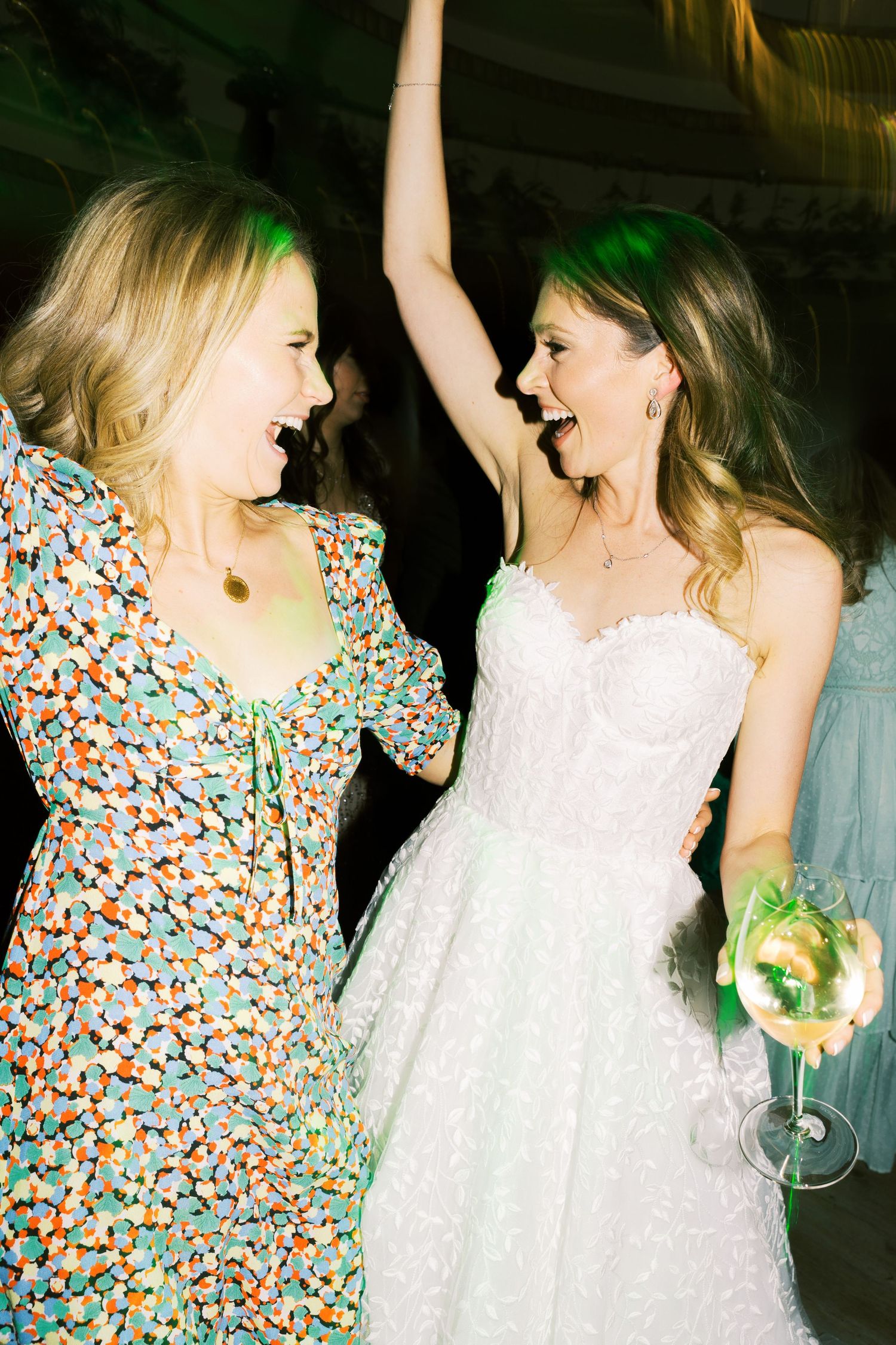 Two women dancing joyfully at a wedding reception, one in a white bridal gown holding a champagne glass.