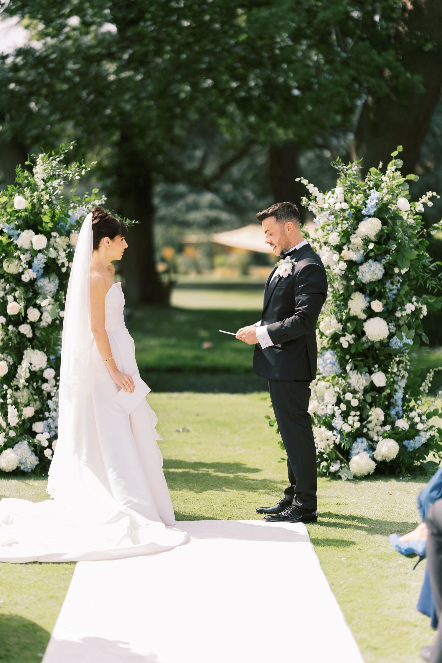 Bride and groom exchange vows outdoors at elegant wedding ceremony with white floral arch and lush greenery backdrop.