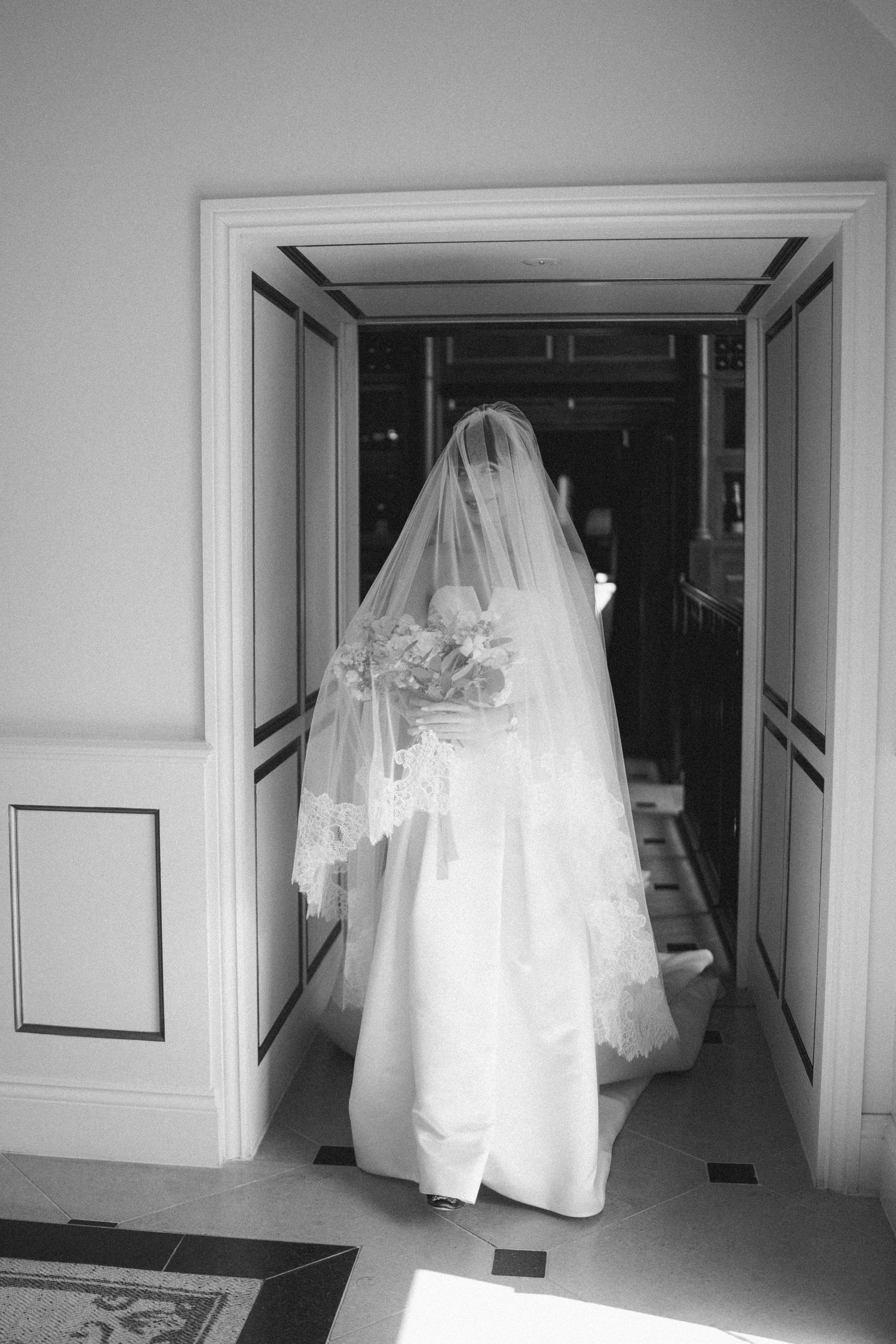 Black and white photo of a bride in a white gown and lace veil holding a bouquet, standing in an elegant doorway.