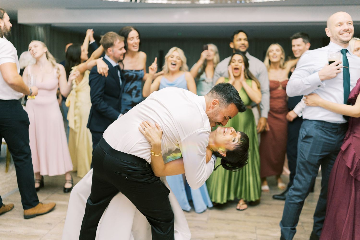 Groom dips bride for a kiss on the dance floor surrounded by cheering wedding guests in elegant attire.