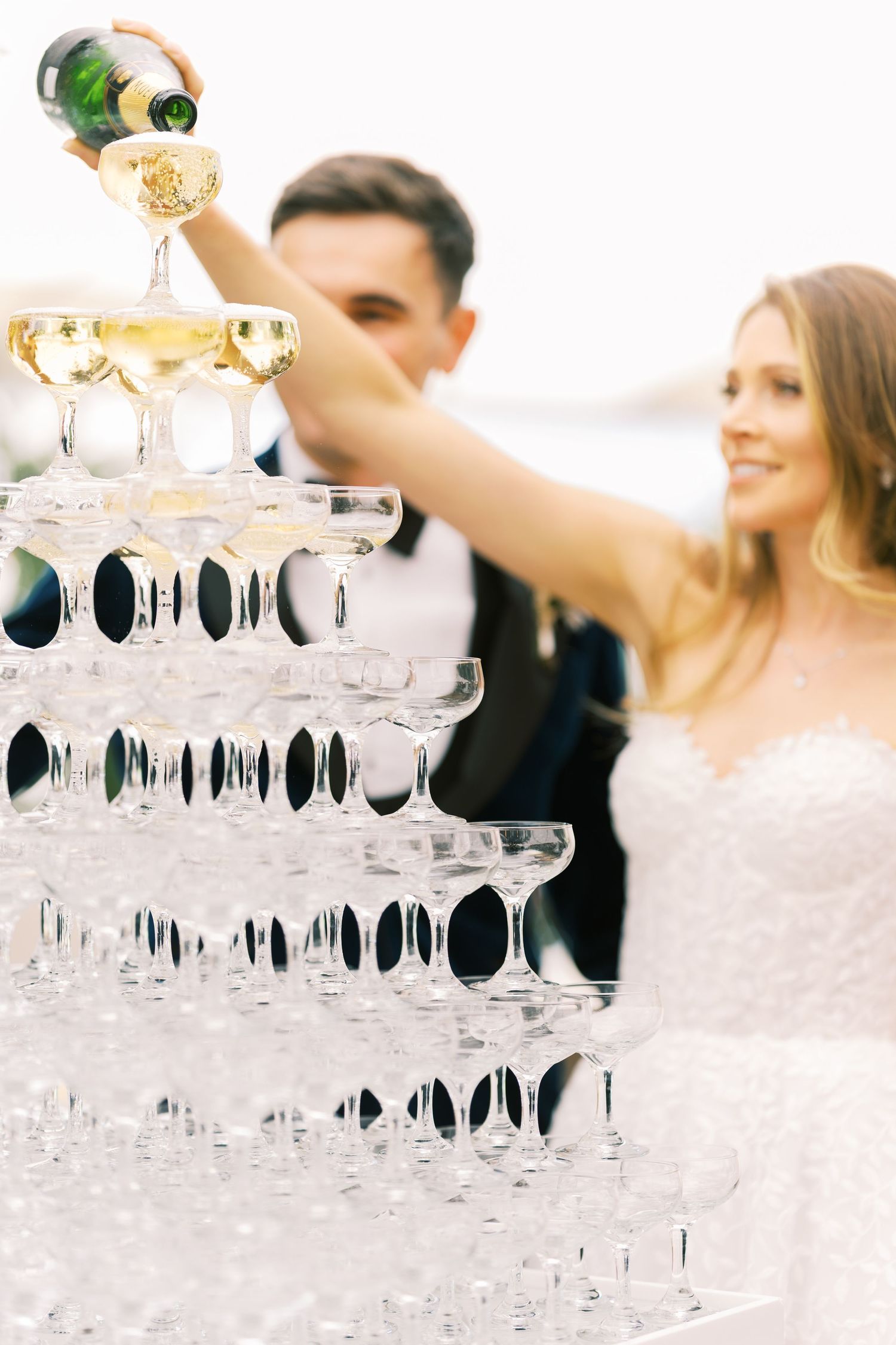Bride and groom pouring champagne into a tall pyramid of glasses at their outdoor wedding celebration.