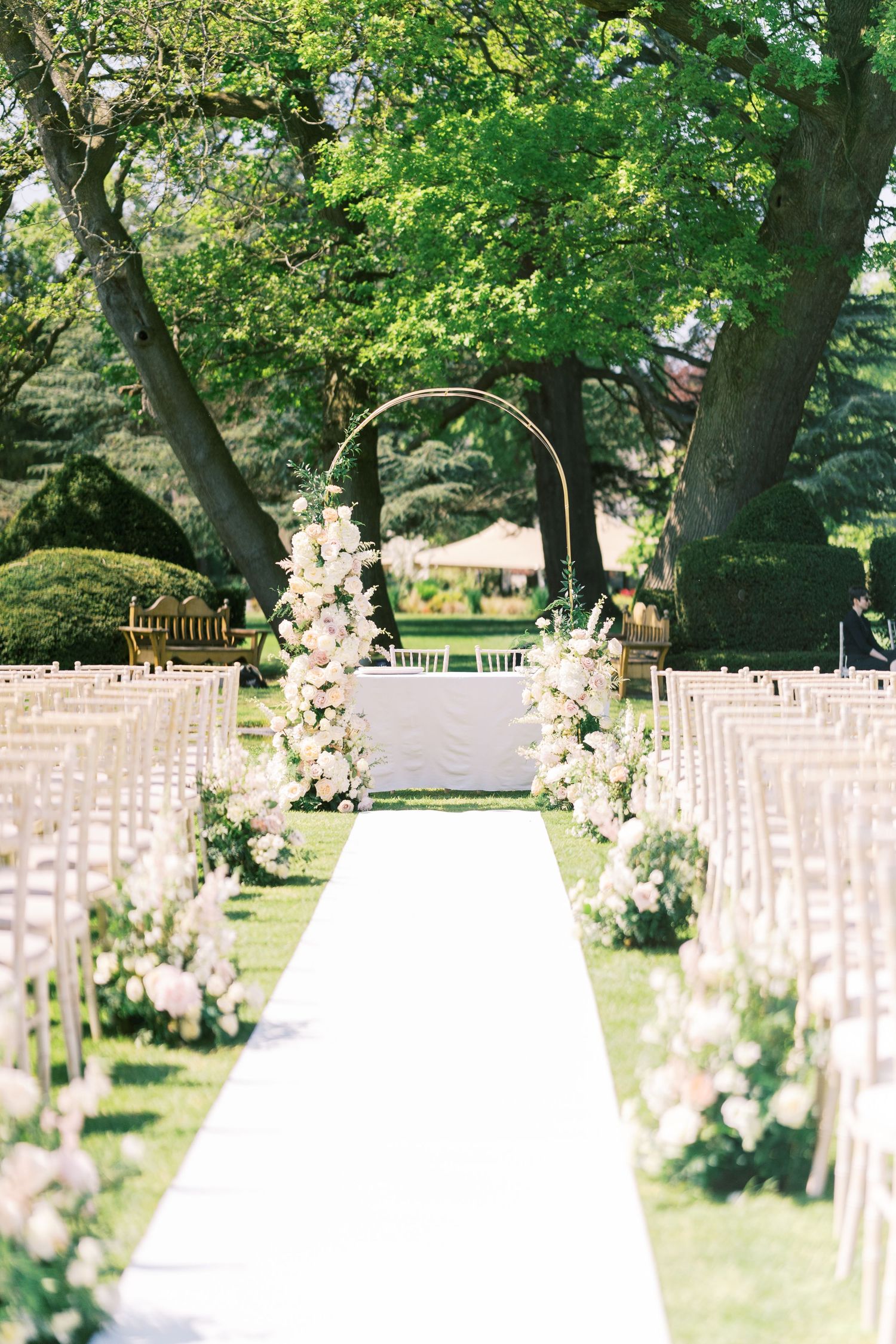 White aisle runner leads to a floral circular arch flanked by lush blooms at an outdoor garden wedding ceremony.