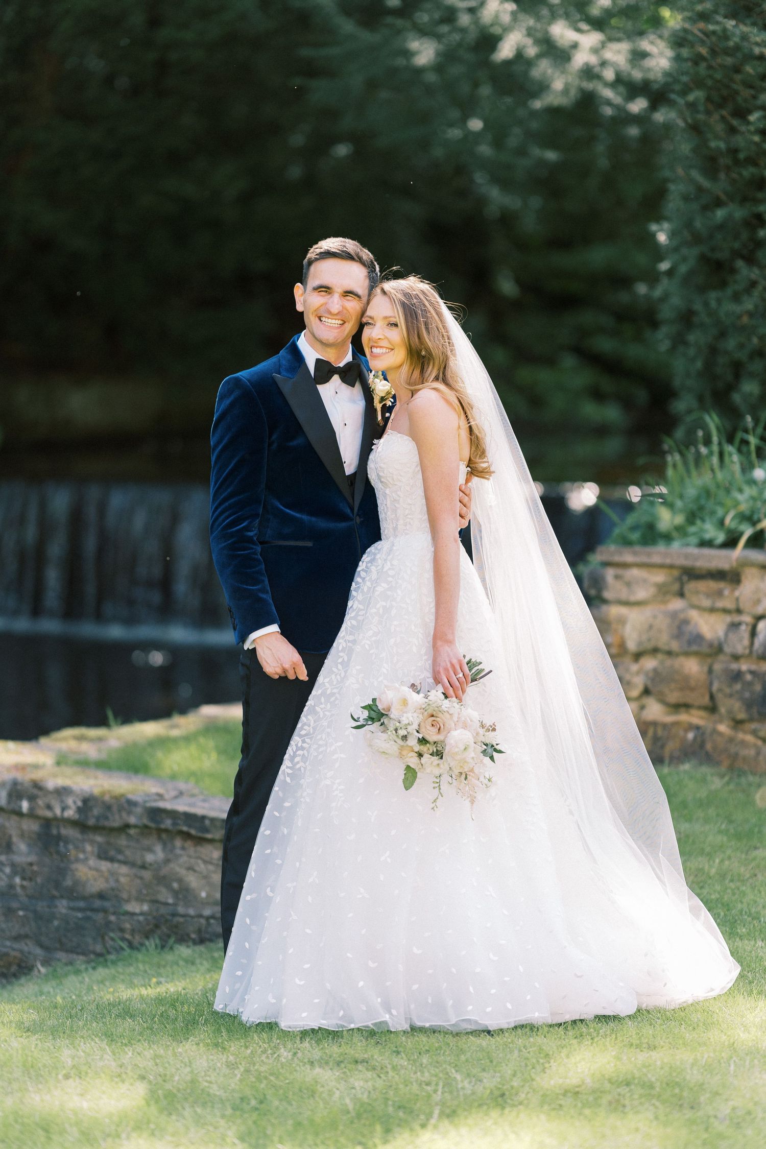 A bride in a white ball gown and groom in navy tuxedo pose together outdoors near a stone waterfall.